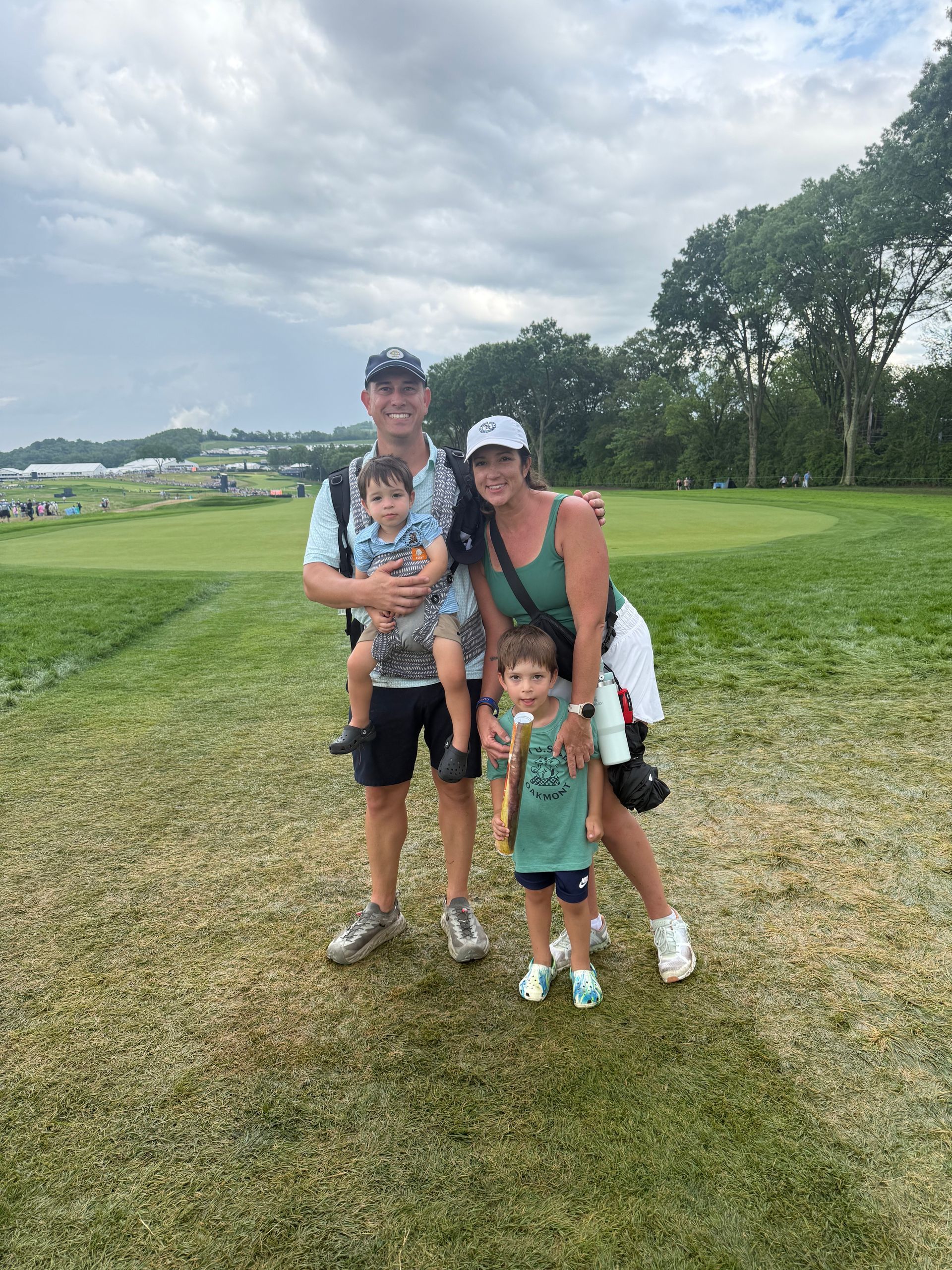 Family of four poses on a golf course. Father carries a child, mother stands with another child. Green clothing.