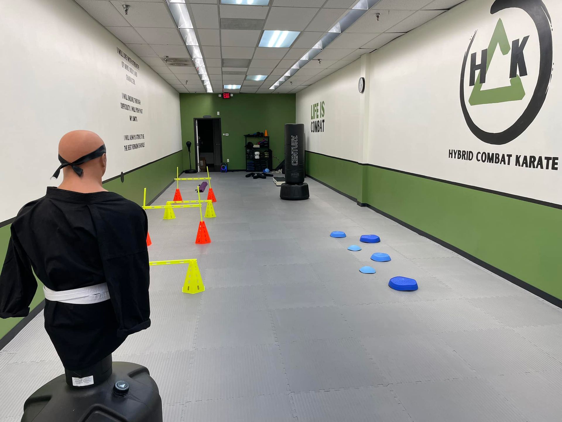 A man in a black karate uniform is standing in a gym with a hk logo on the wall.