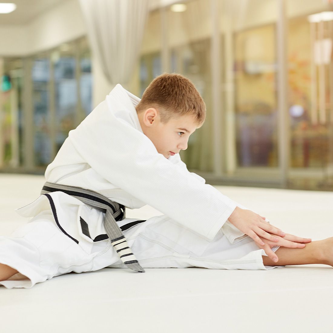 A young boy in a karate uniform is sitting on the floor stretching his legs.