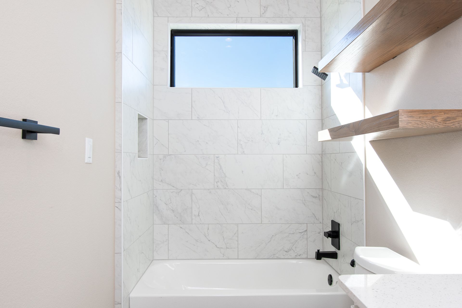 A bathroom with a tub , toilet , shelves and a window.