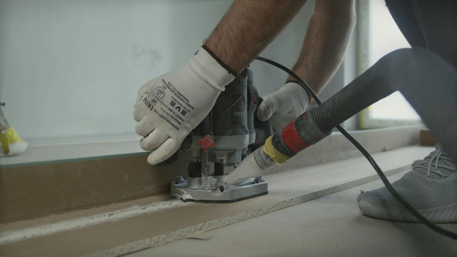 Person using a router tool on a wooden surface, wearing work gloves, with a vacuum hose attached.