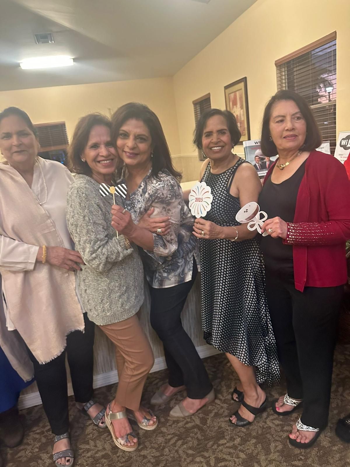 Five women posing together indoors; holding props. Some wear dresses, cardigans.