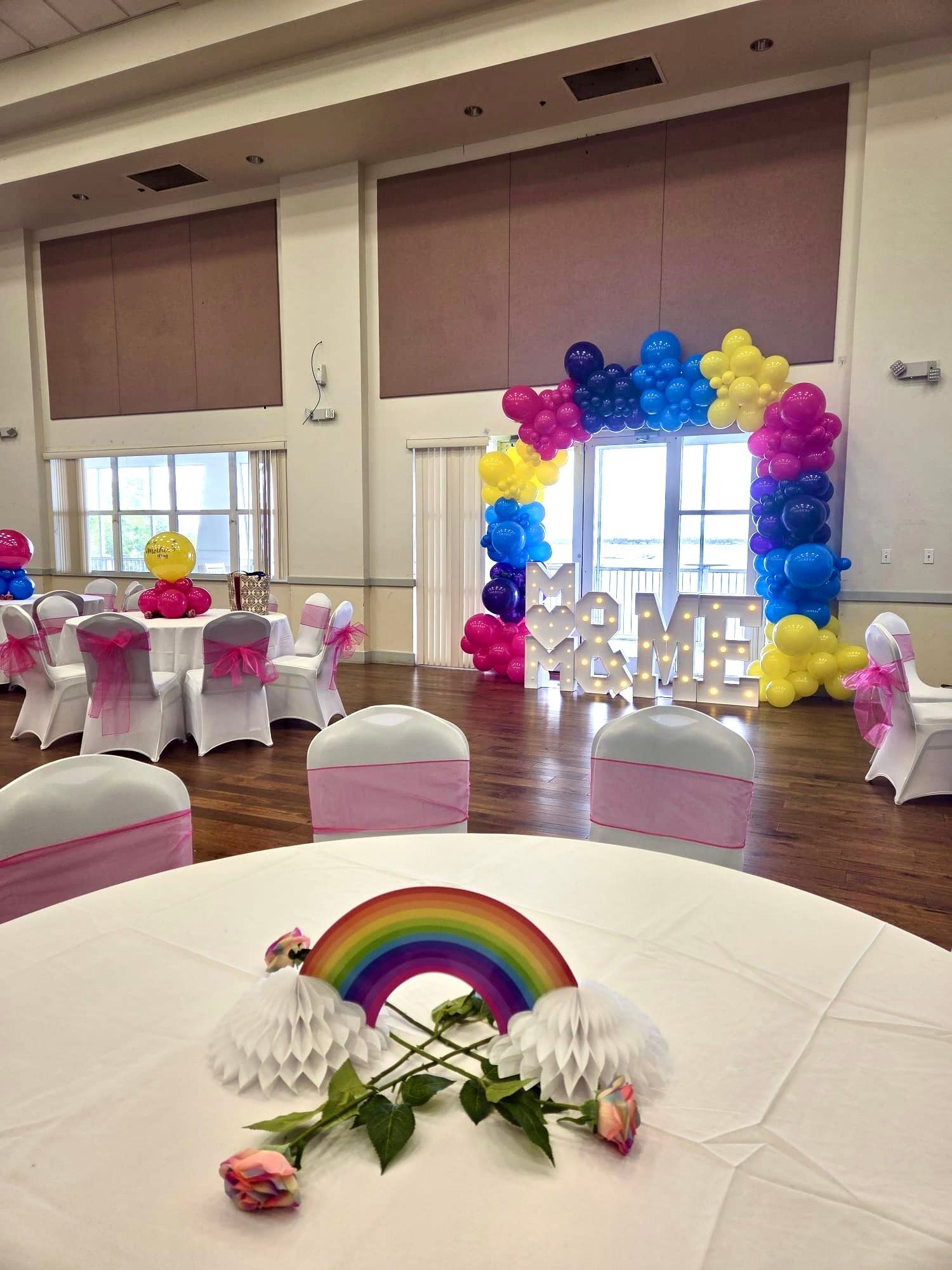 Party venue decorated with rainbow-themed balloons and table centerpieces. White chairs with pink accents.