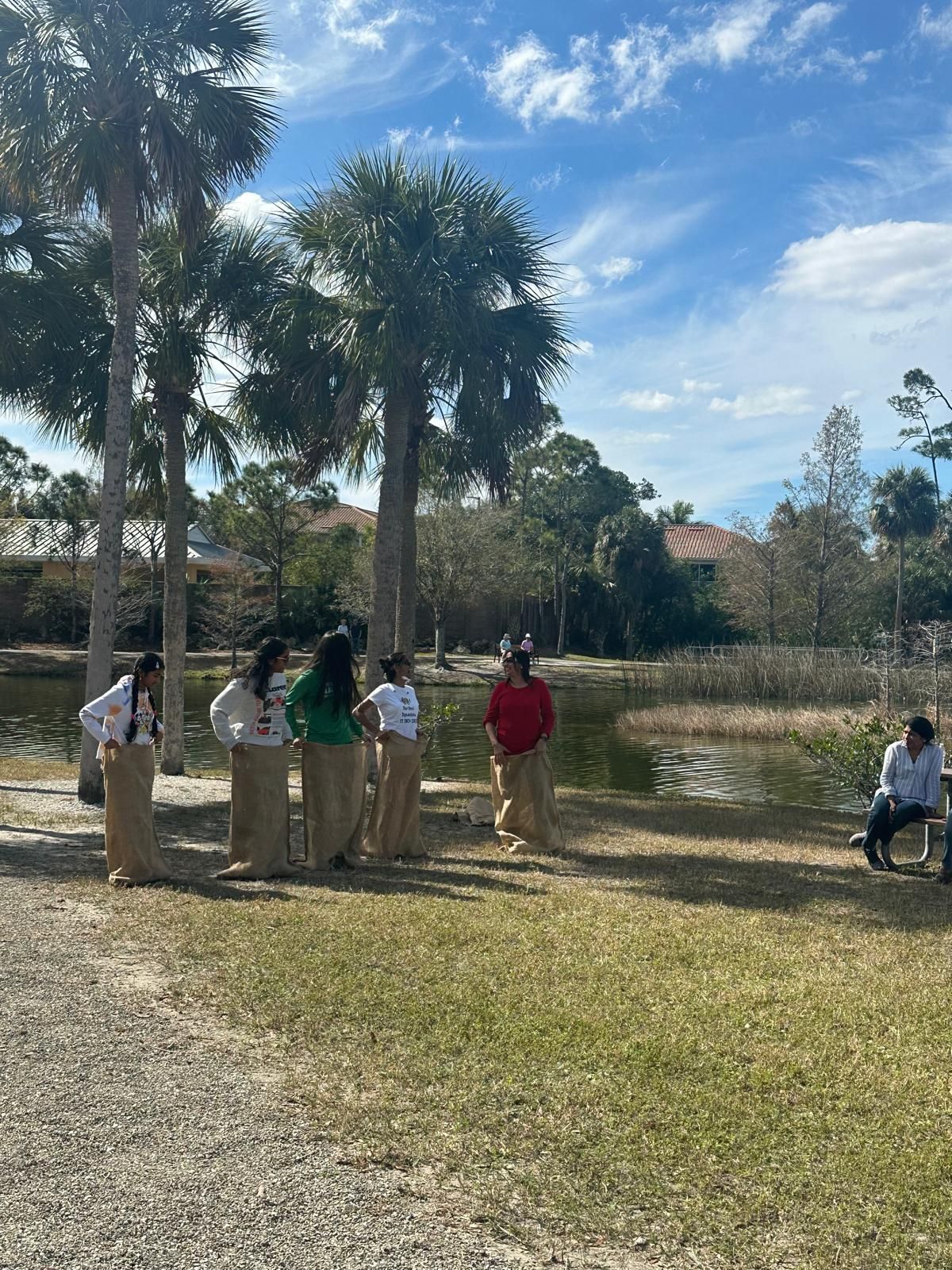 People in sacks ready to race on grass, with trees and a pond in background. Bright sunny day.