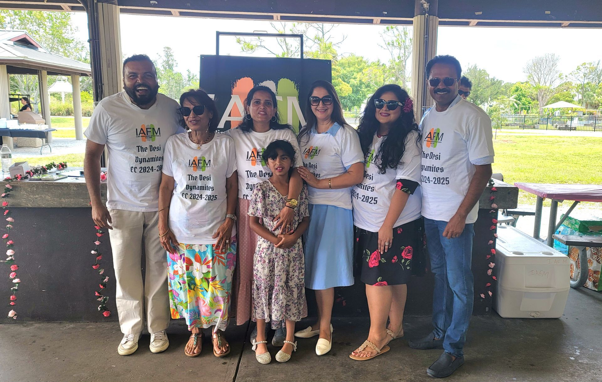 Group of people in white t-shirts posing outside. They stand near a black sign, smiling.