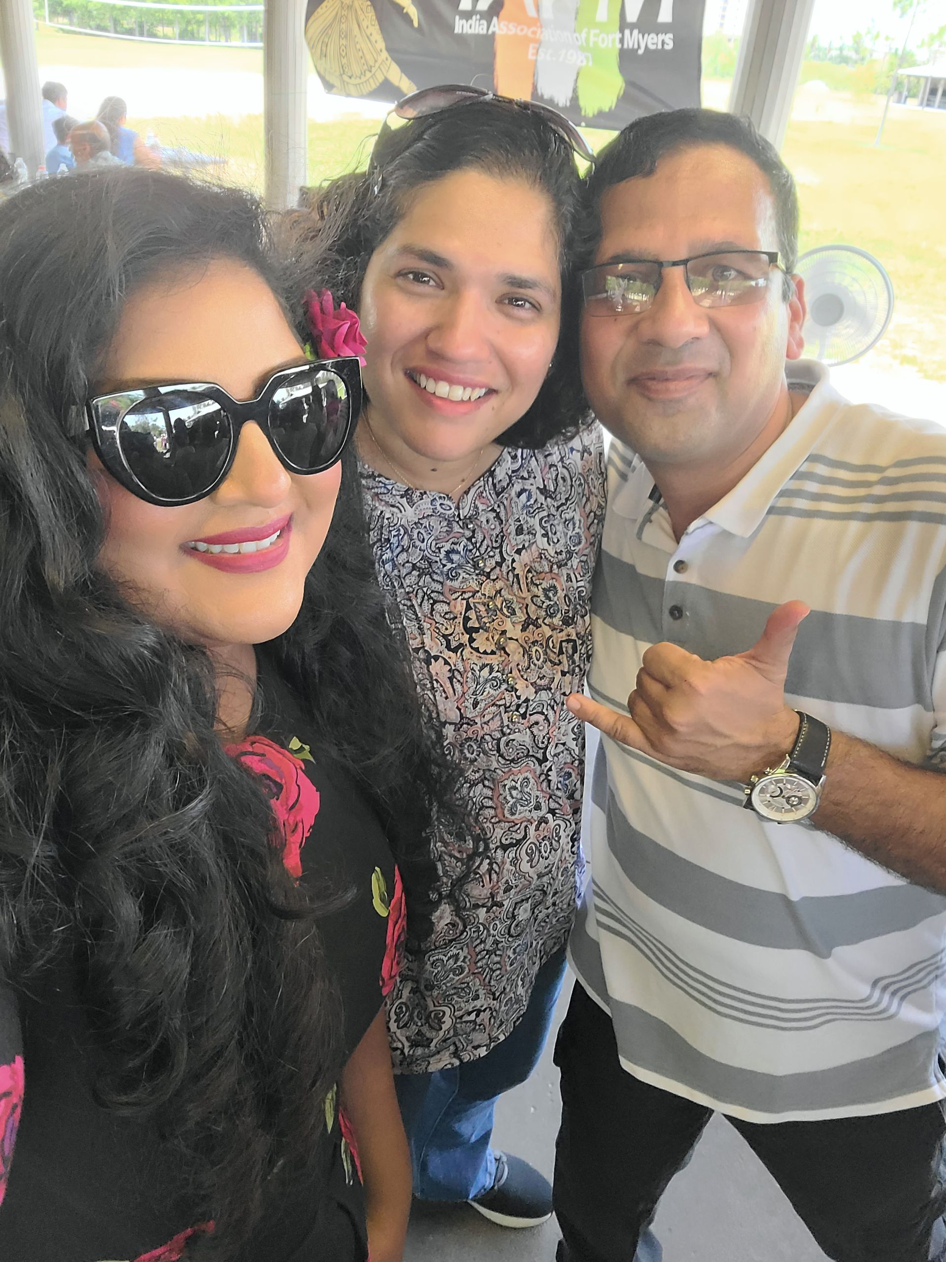 Three people smiling for a selfie. Woman on left wearing large sunglasses, man on right giving shaka sign.
