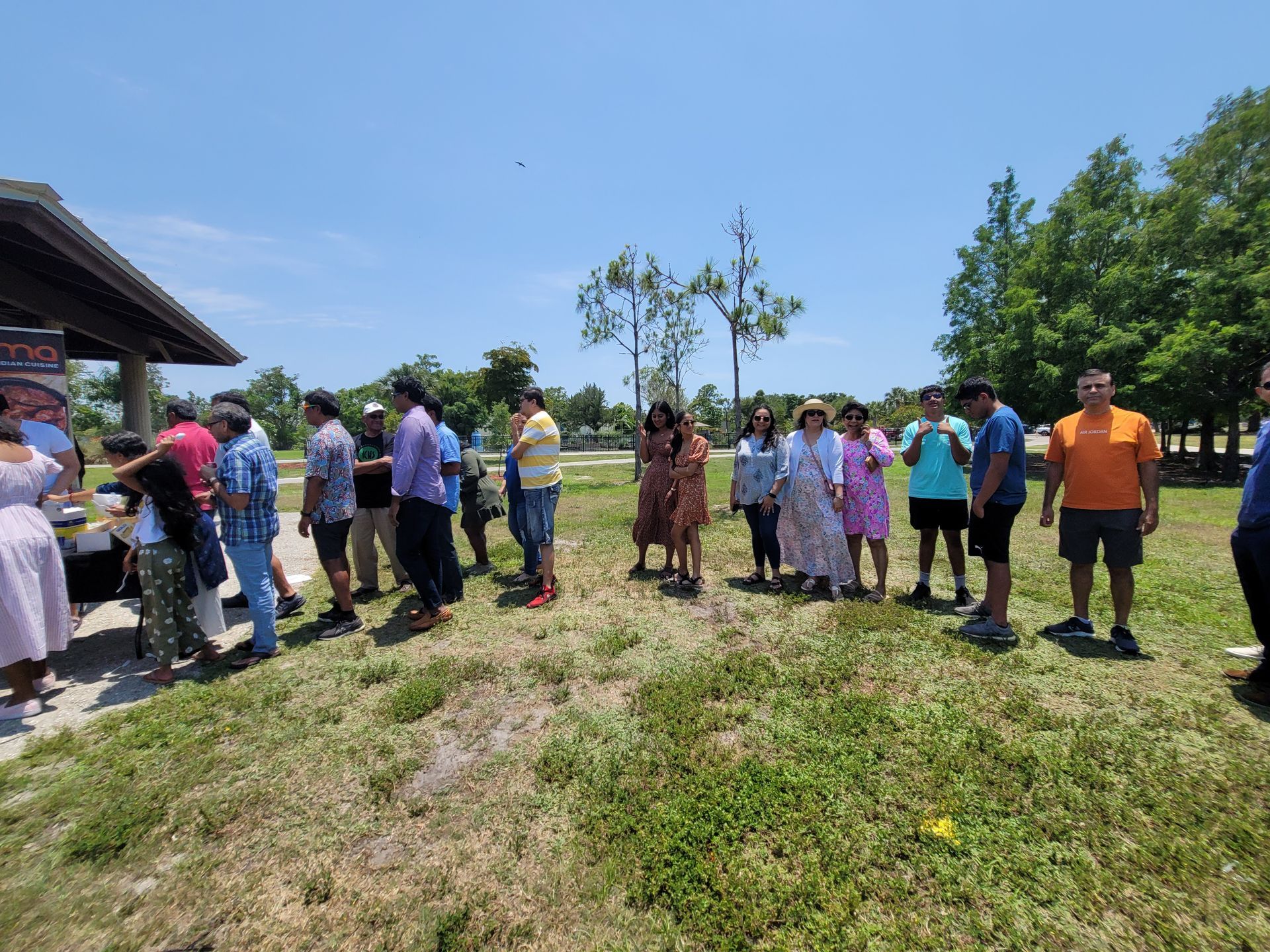 People gathered outdoors, waiting in line under a pavilion on a sunny day.