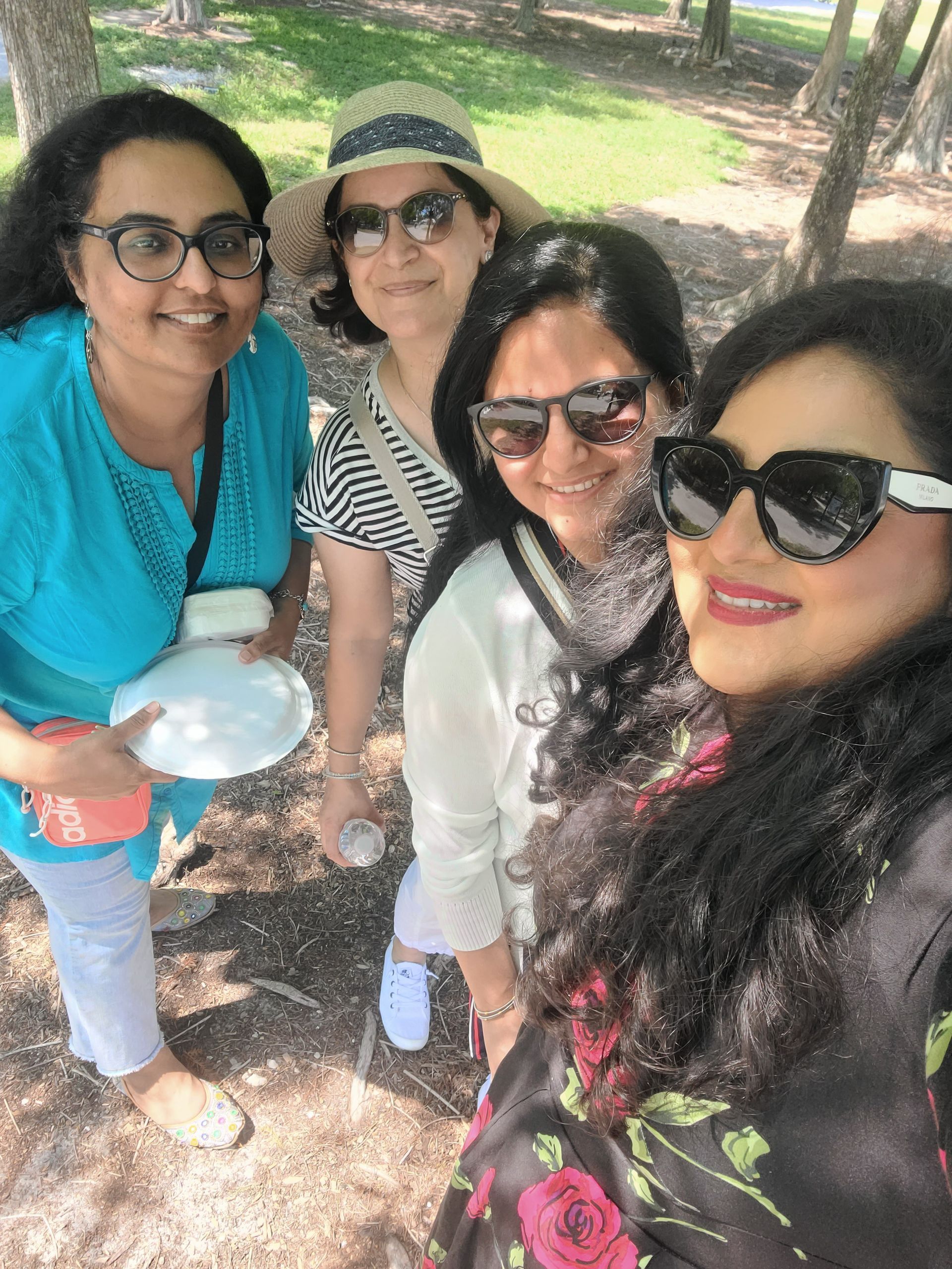 Four women smiling, taking a selfie outdoors. Sunlight, one in blue, one in hat, one in white, one in floral.