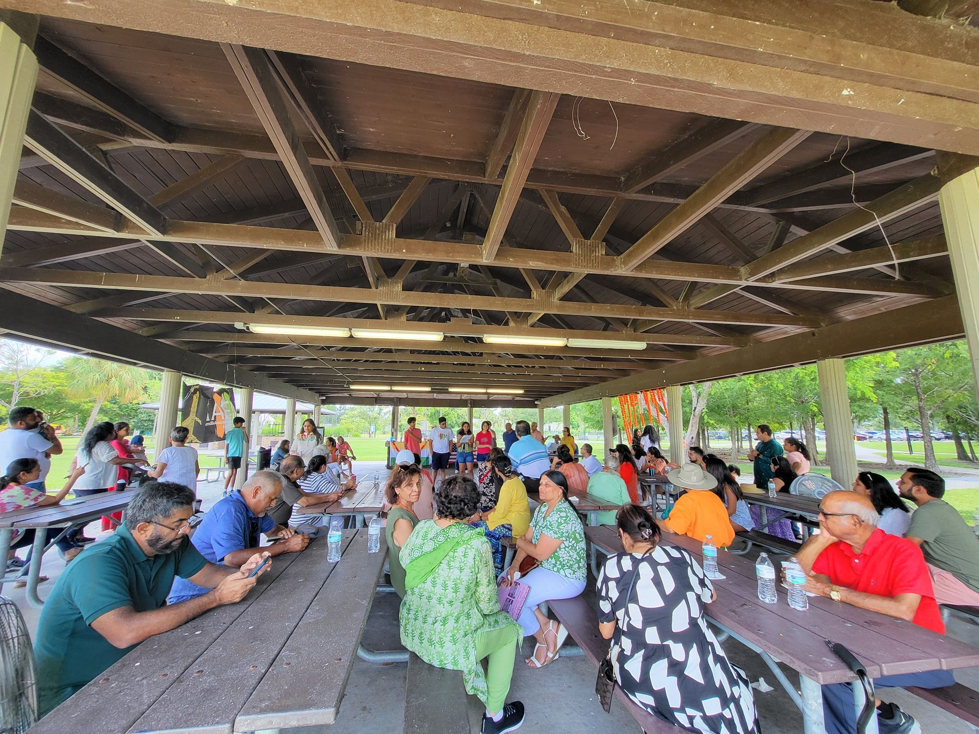 Large group of people under a covered picnic area, socializing. Sunny outdoor setting.