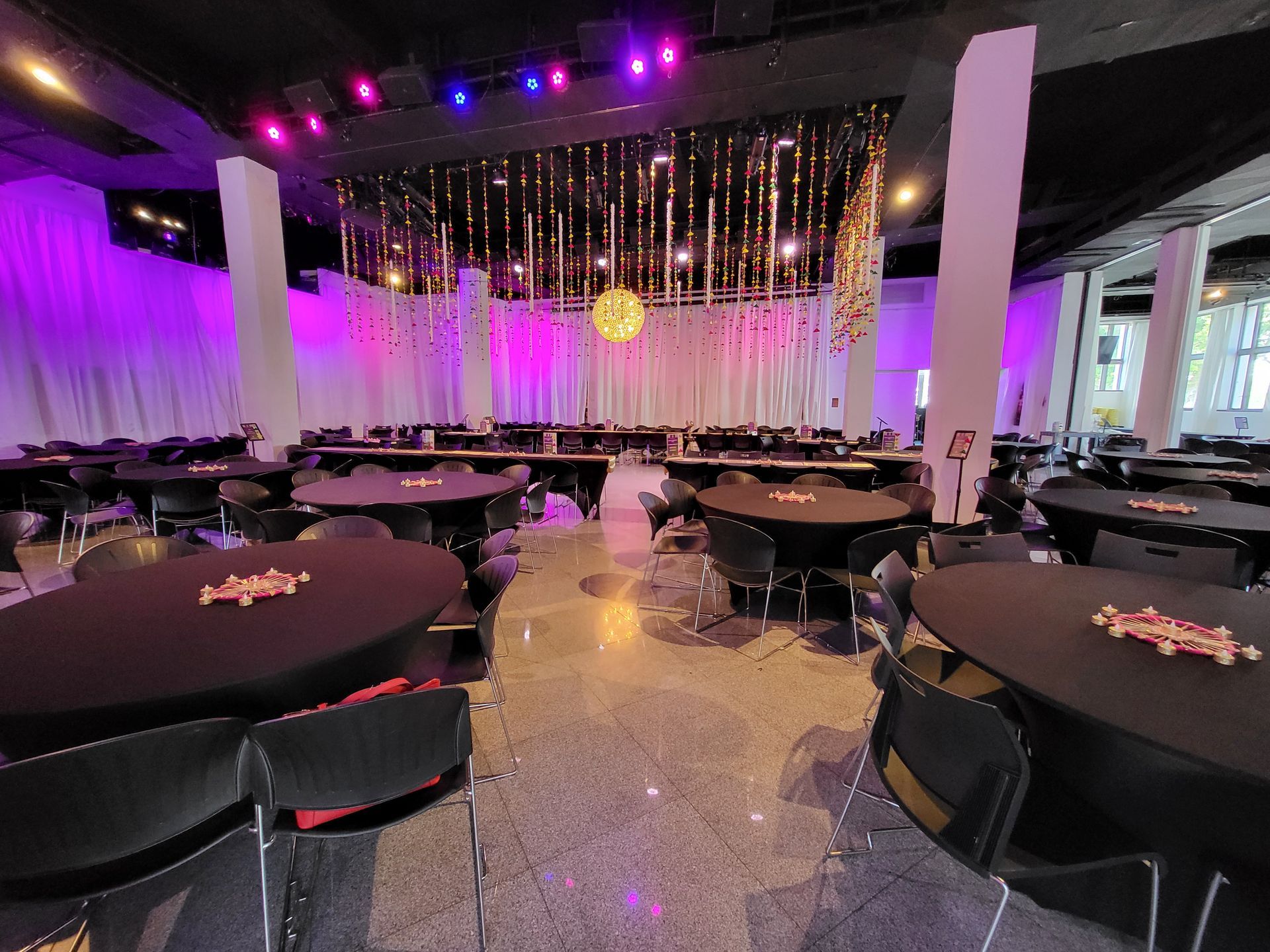 Event hall with round tables covered in black linens. Purple and gold lighting accents a decorated stage.