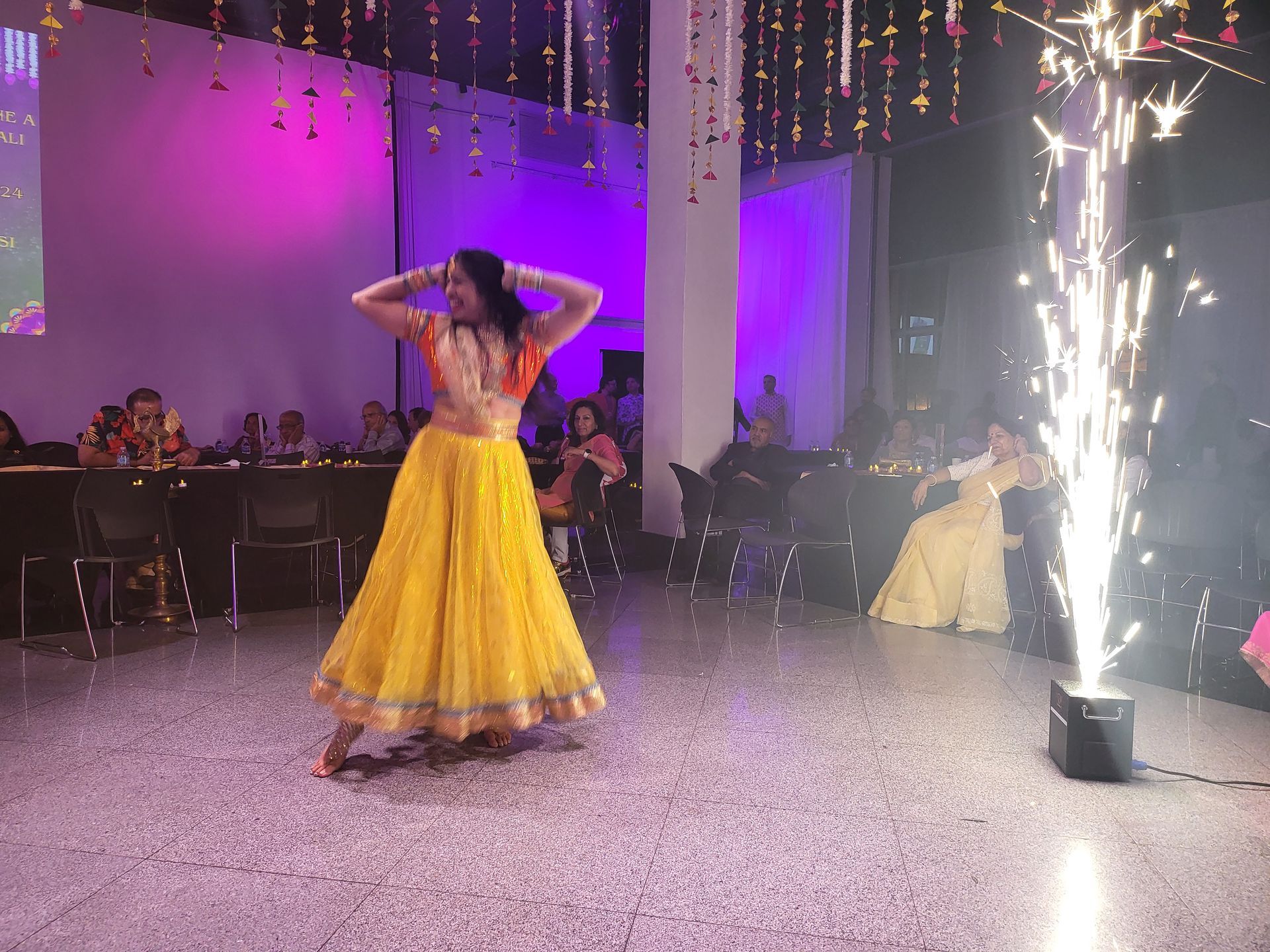 Woman in yellow and orange outfit dancing with sparkler effect at an event.
