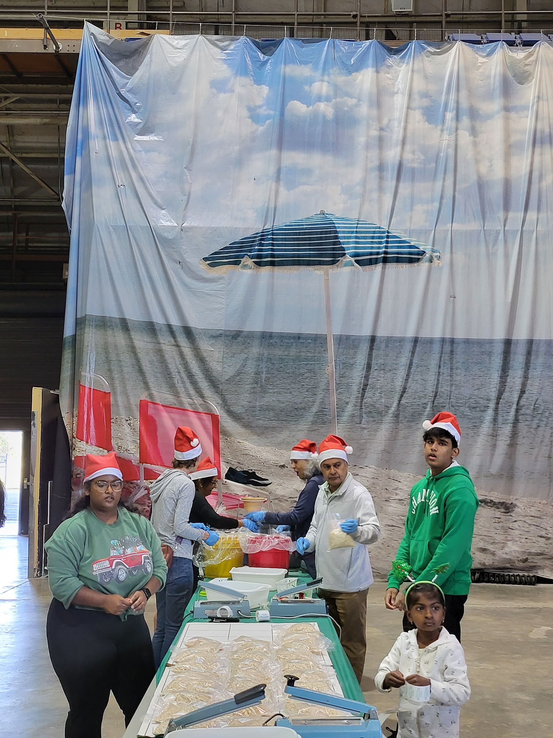 People in Santa hats at a beach-themed event. Table set up, large beach scene backdrop.