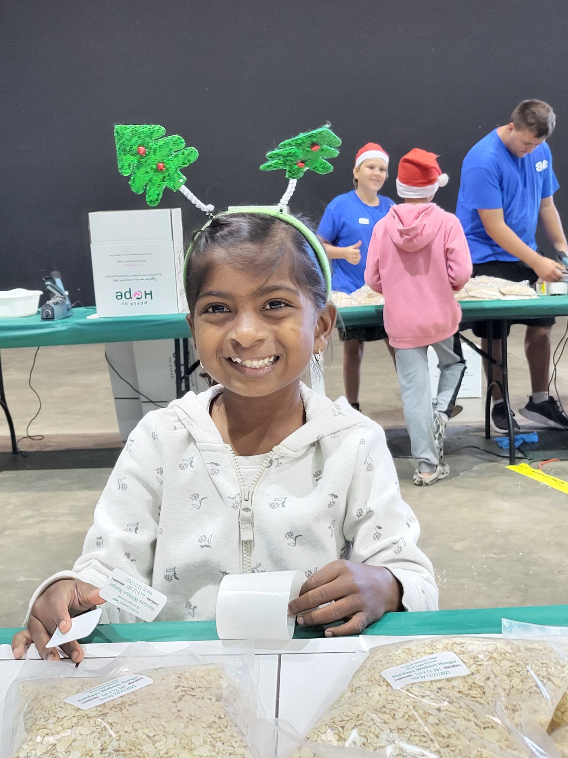 Girl with Christmas tree antlers smiles at a table; others pack bags behind her.