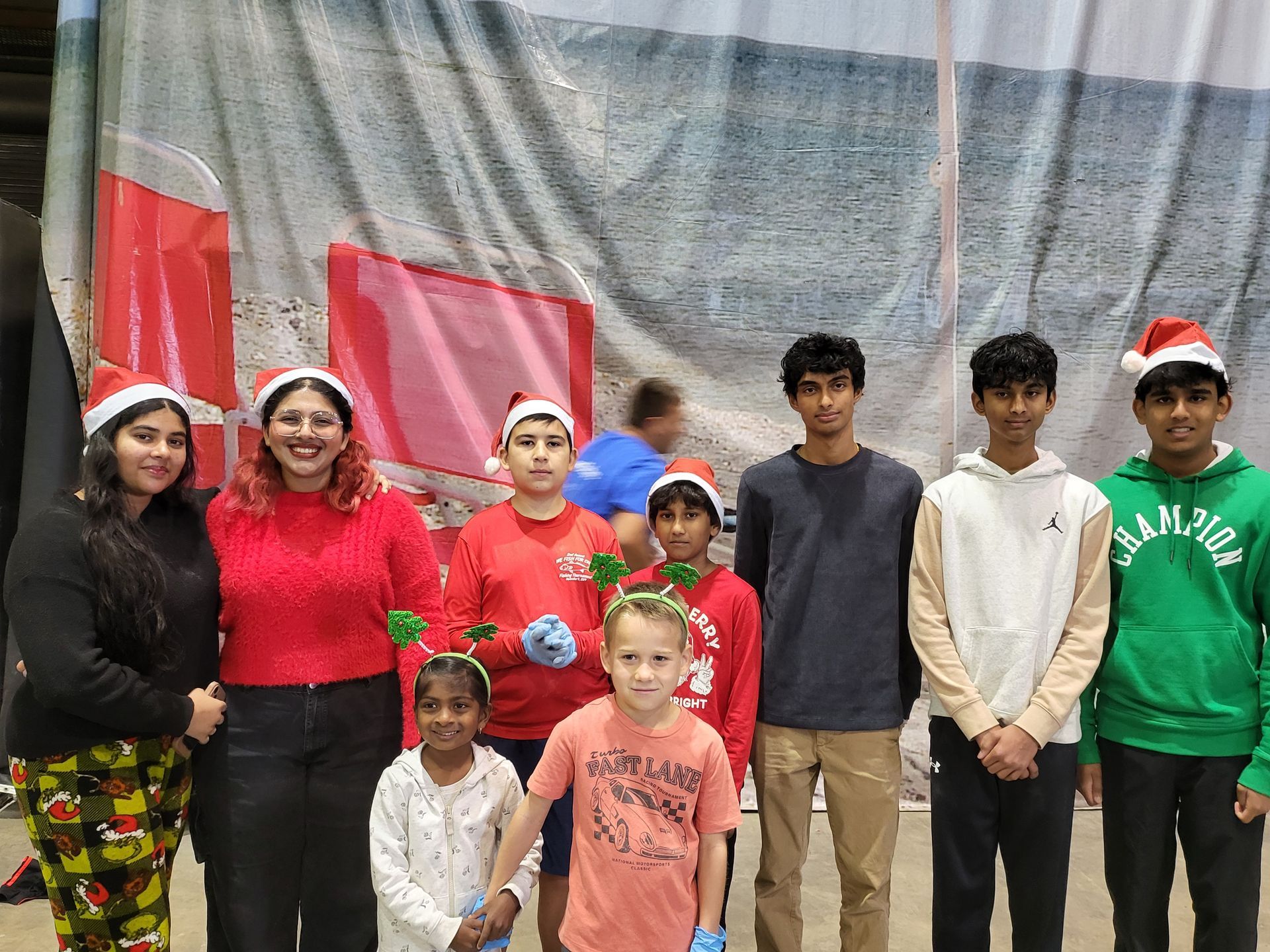 Group of people wearing Santa hats, standing in front of a backdrop; some children hold small blue items.