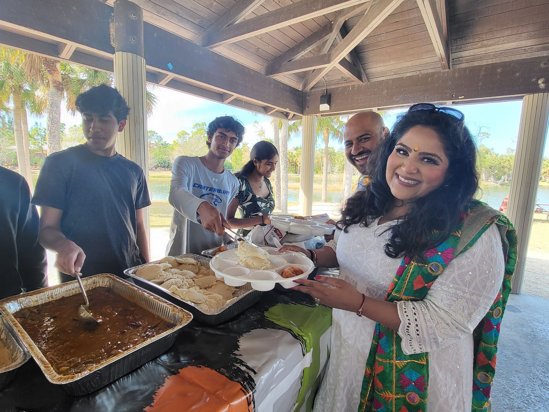 Family serves Indian food at an outdoor gathering under a pavilion.