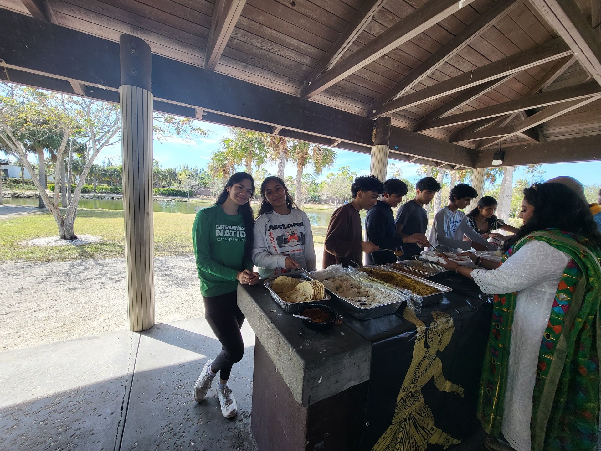 People at an outdoor gathering with food. Women and men smiling, sunshine, green grass, shelter with brown posts.