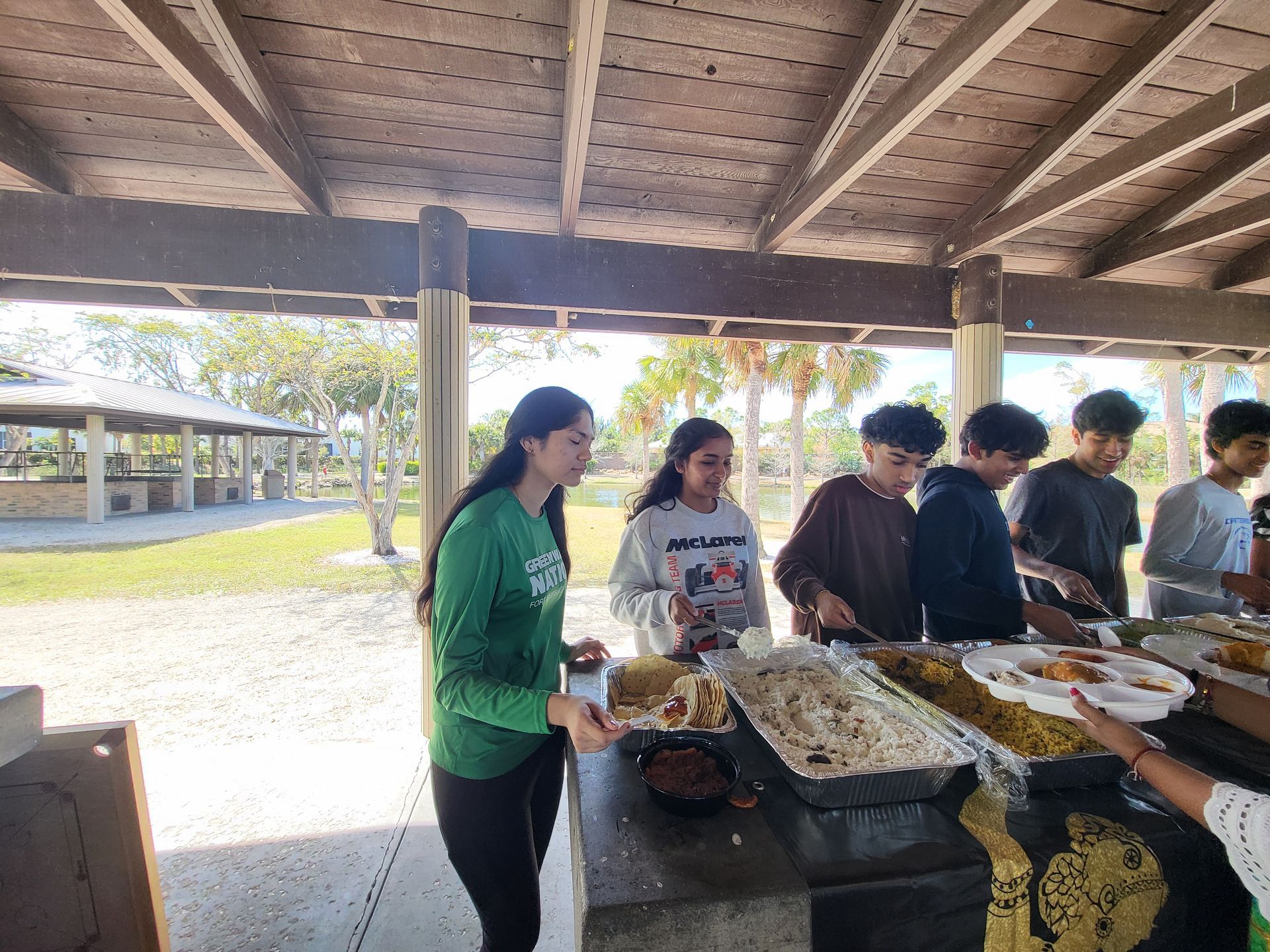 Group of people serving food at a picnic table in a park under a covered area.