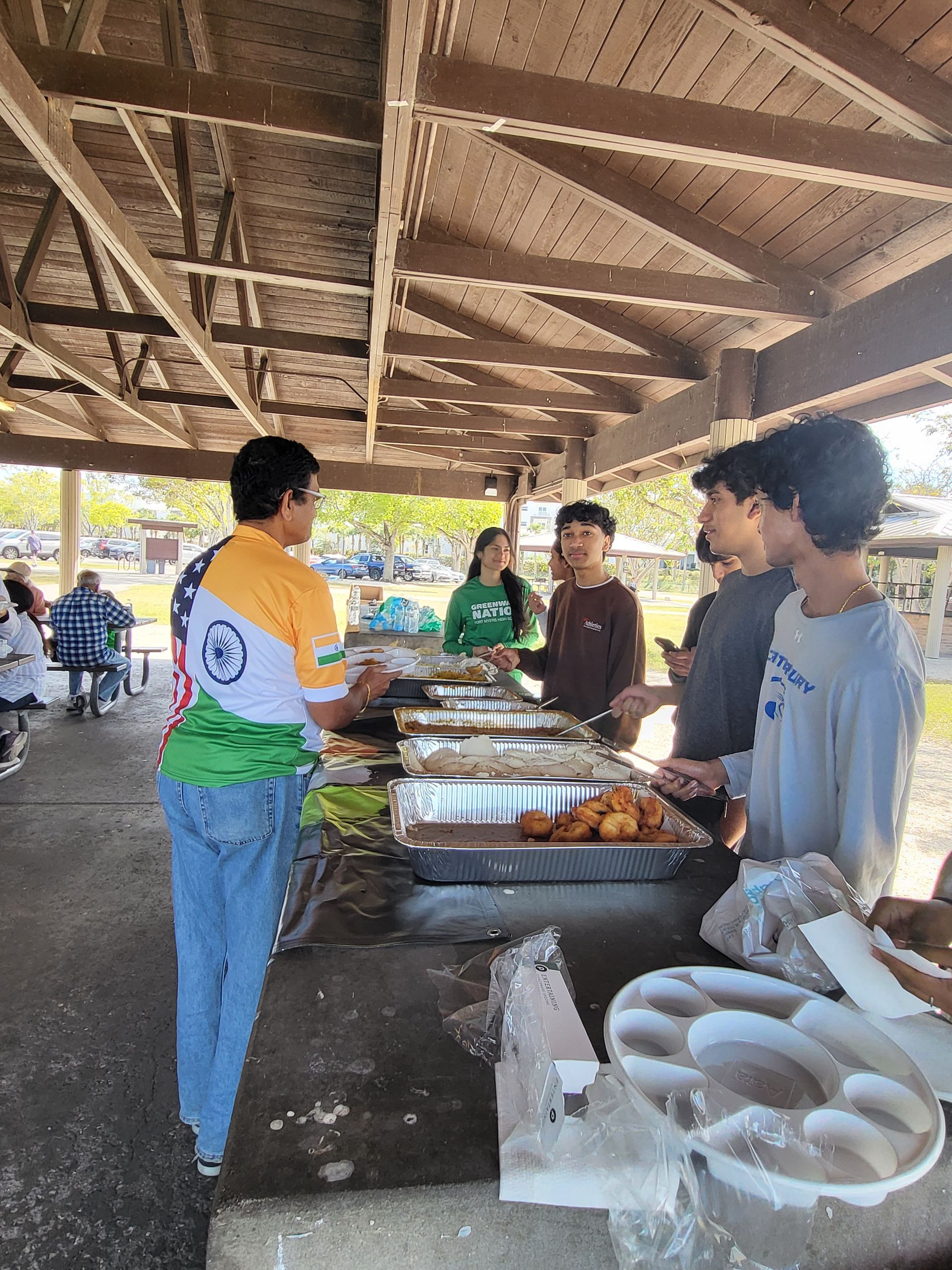 People at a picnic under a shelter with food on tables. One person wears an Indian flag-themed shirt.