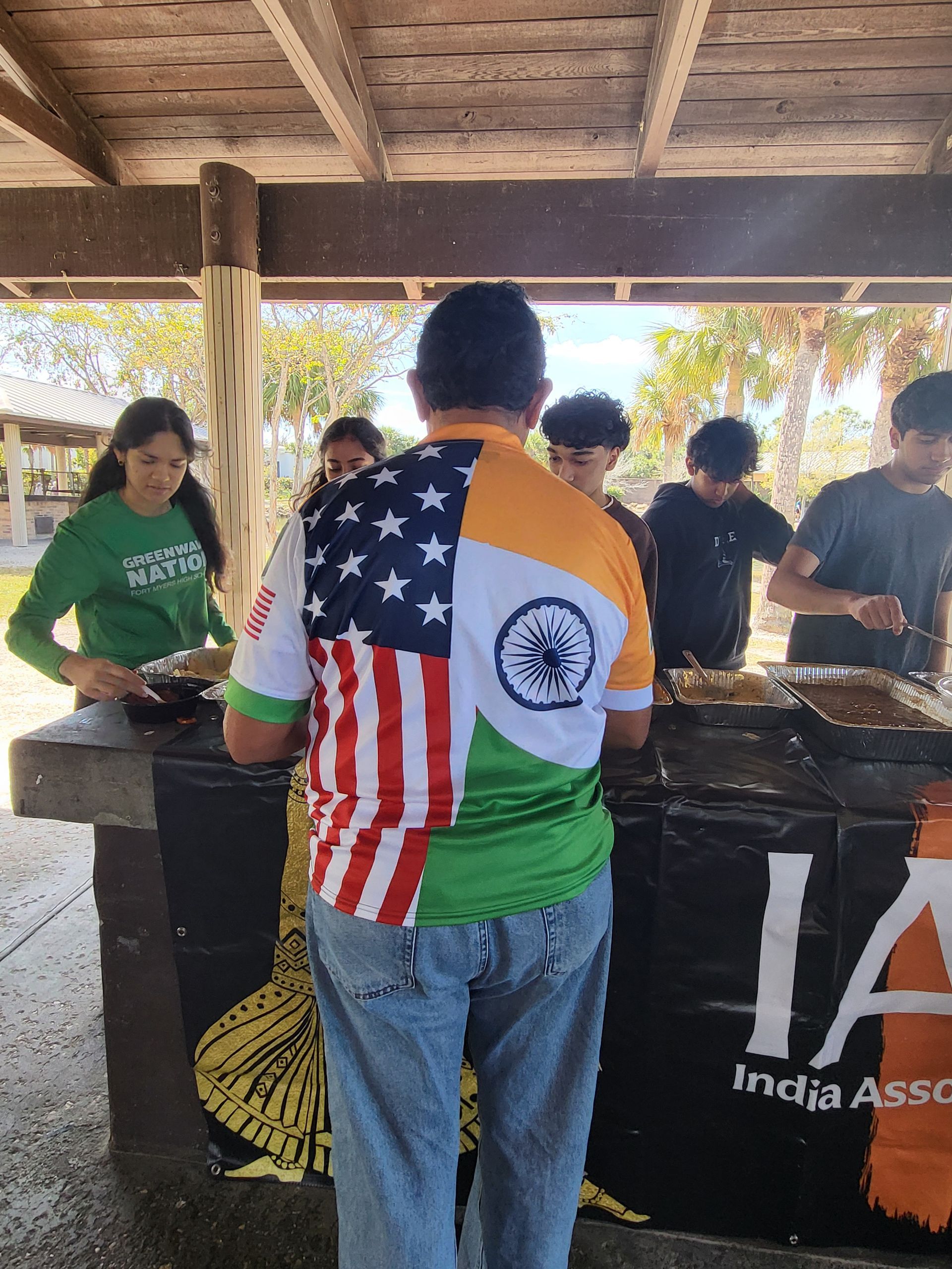 People at an Indian food event. A man in shirt with Indian and American flags, preparing food. Others serving.