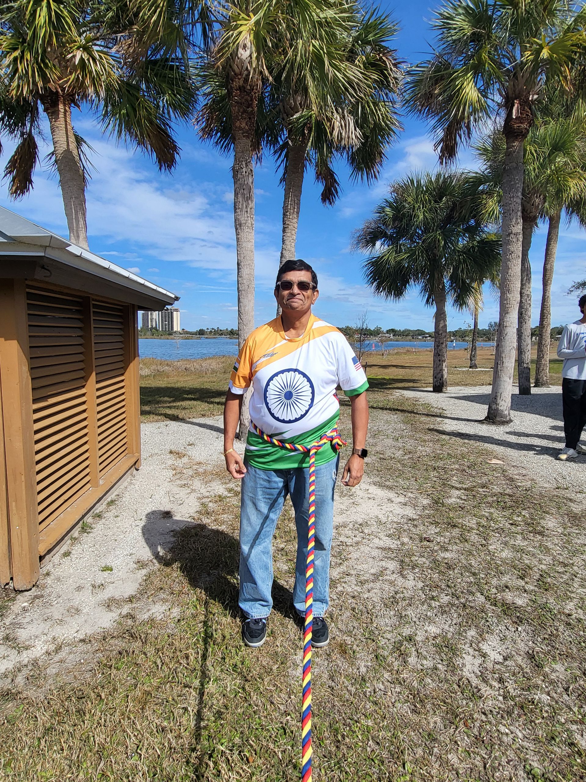 Man in India flag shirt with rope, blue jeans, standing by palm trees and water.