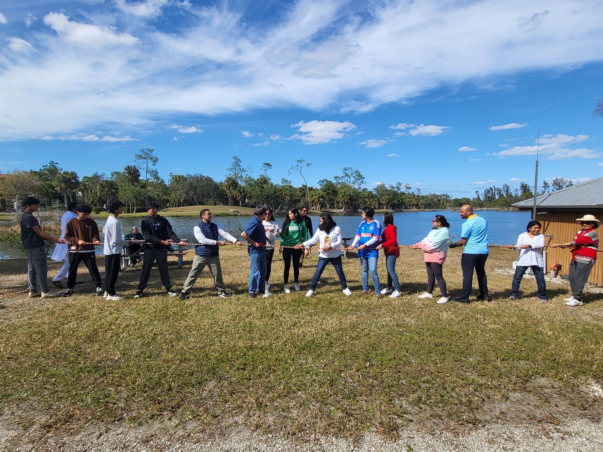 People playing tug-of-war by a lake under a blue sky.