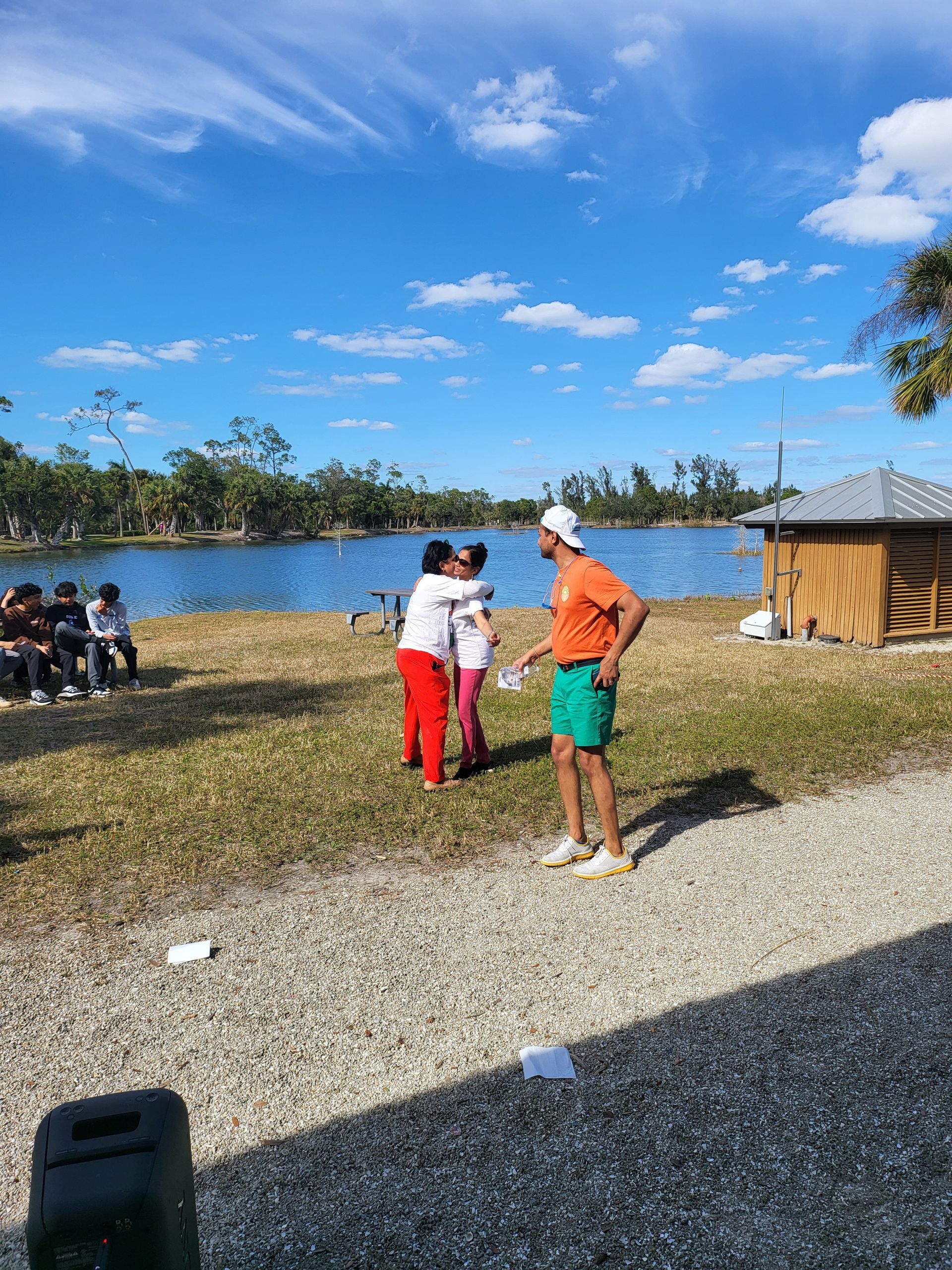 People hugging near a lake, another person in shorts looks on, others sit nearby under a blue sky.