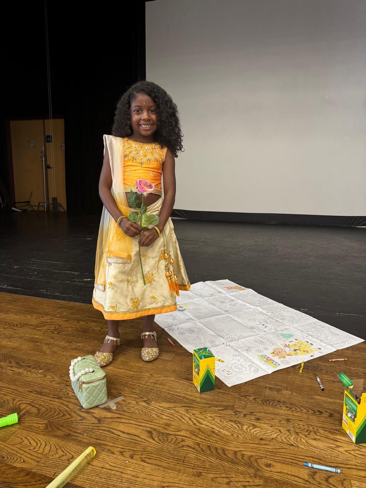 Young girl in yellow dress holding flower smiles, standing on stage near art supplies and large drawing.