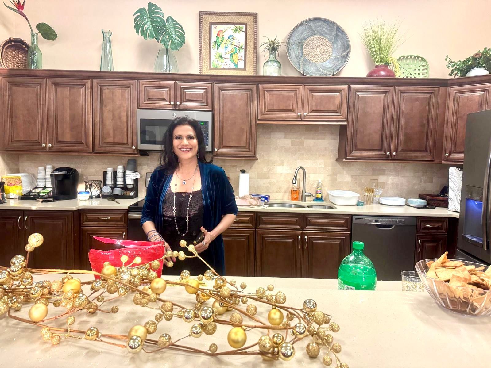 Woman in kitchen smiles, standing near island with gold decor. Brown cabinets, counter, and appliances are visible.