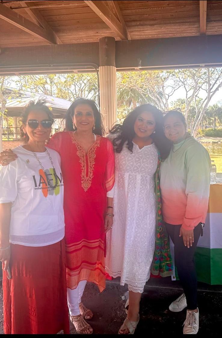 Four women smiling, posing together outdoors. One in white, another in red, others in colorful tops.
