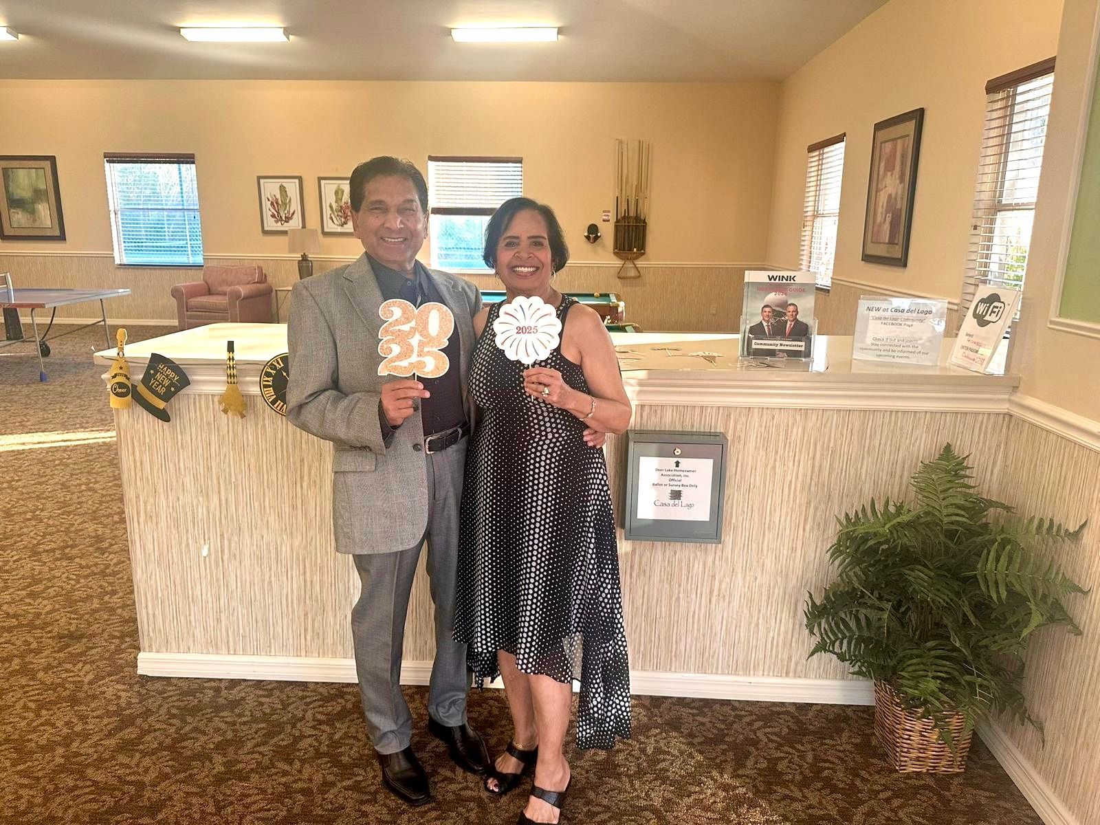 Couple holding cutouts, posing at a party. Man in suit, woman in patterned dress; neutral-toned room.