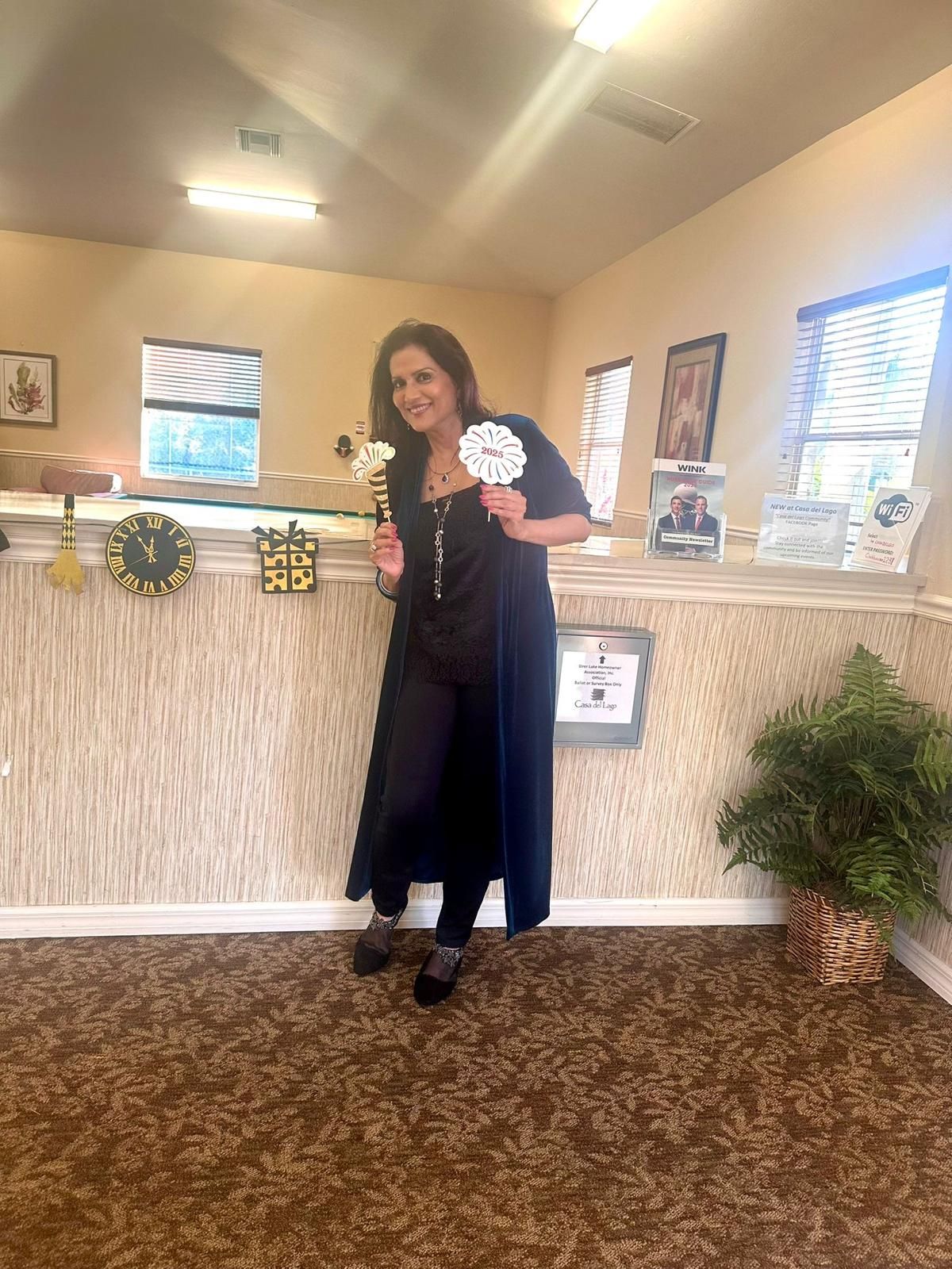 Woman in black, holding a flower and clock, standing near a wall with clocks.