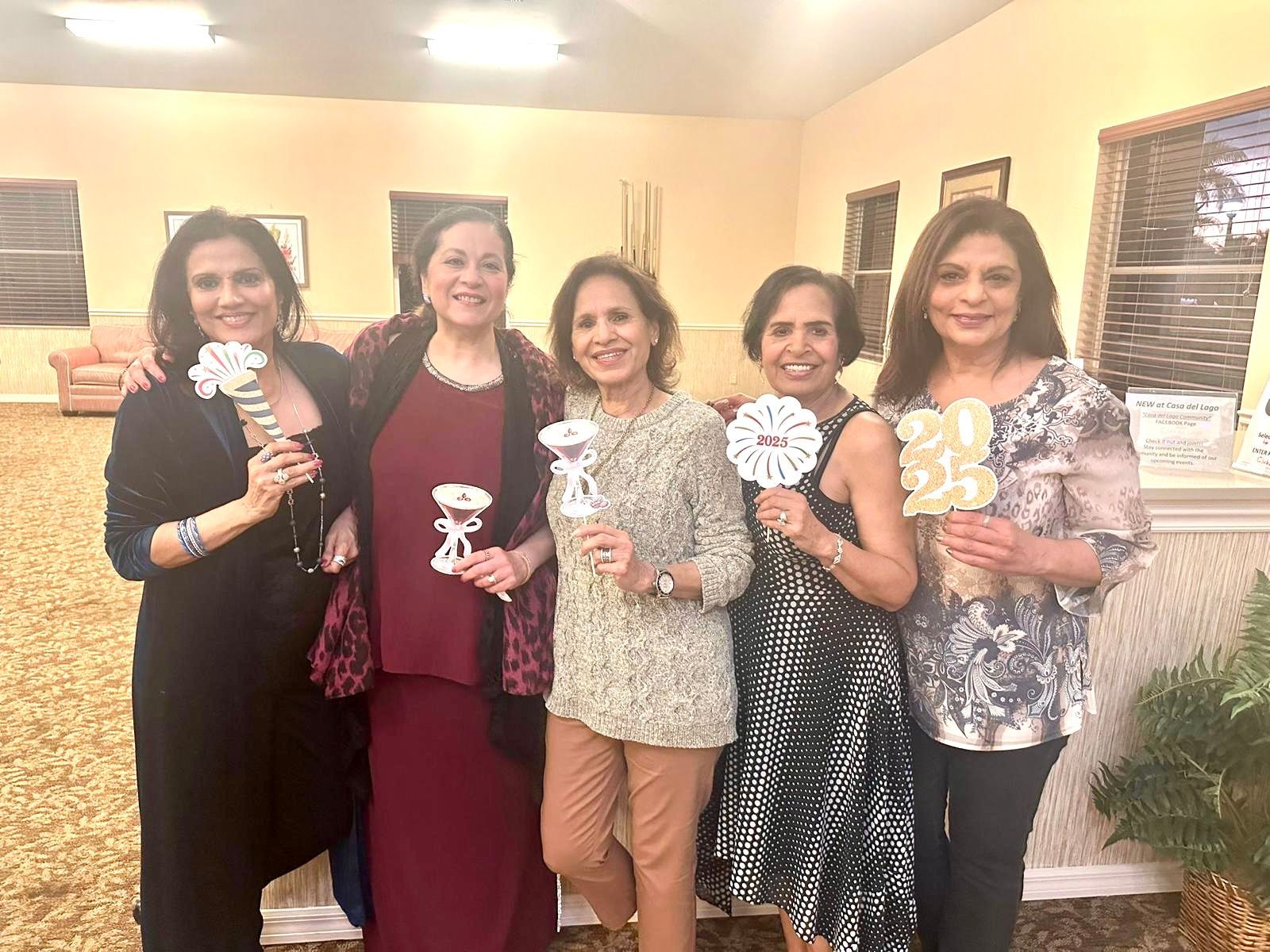 Five women smiling, holding floral decorations, posing indoors at a party.