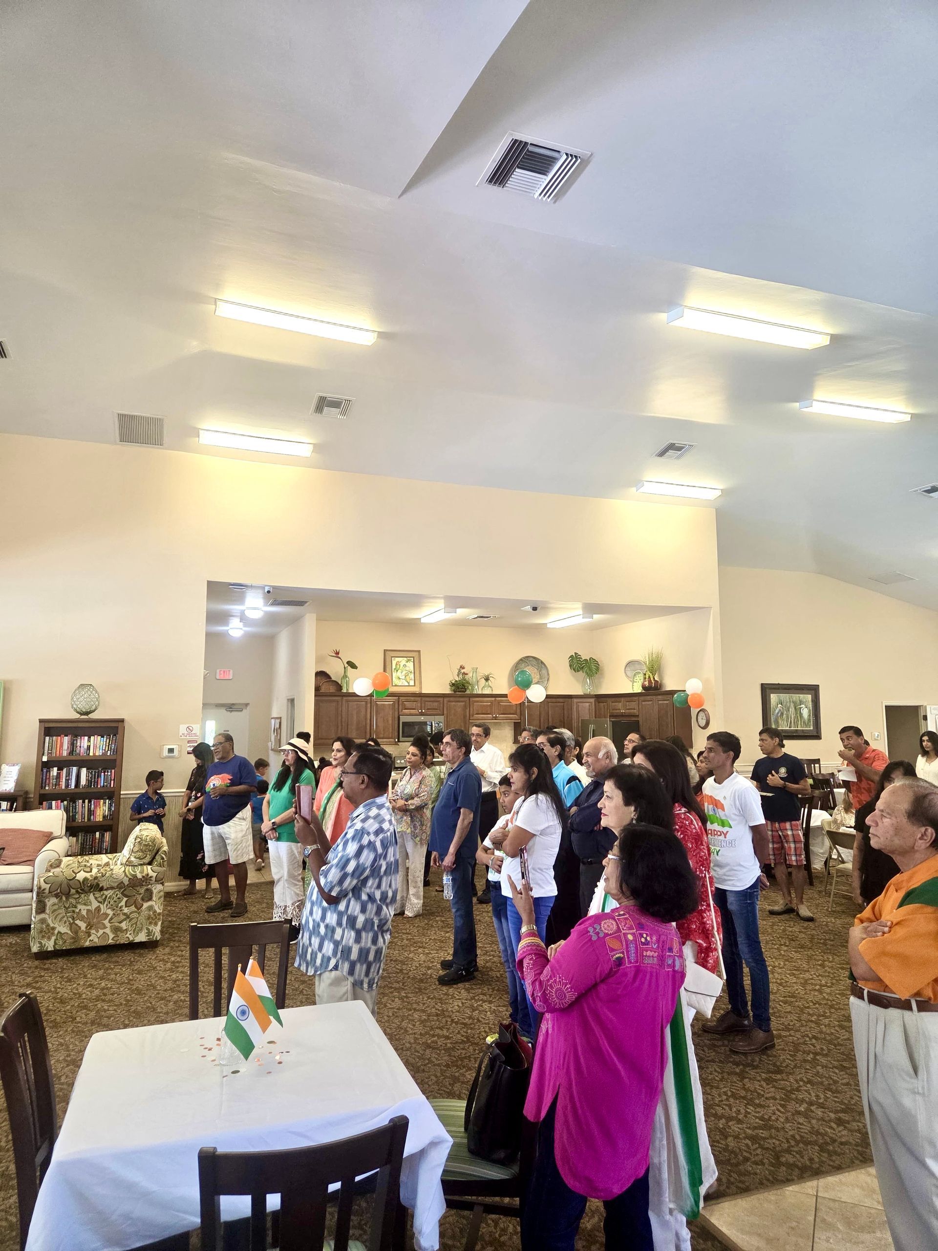 People standing in a room, possibly an Indian Independence Day celebration. Some hold hands over their hearts, tables with flags.