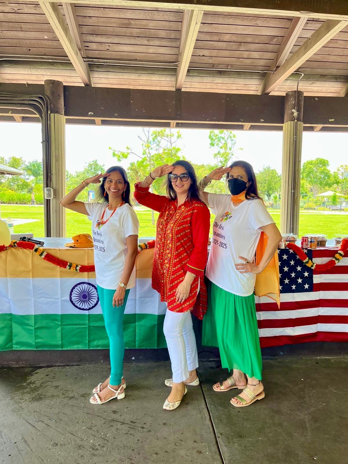 Three women in front of an Indian flag salute with smiles, outdoors.