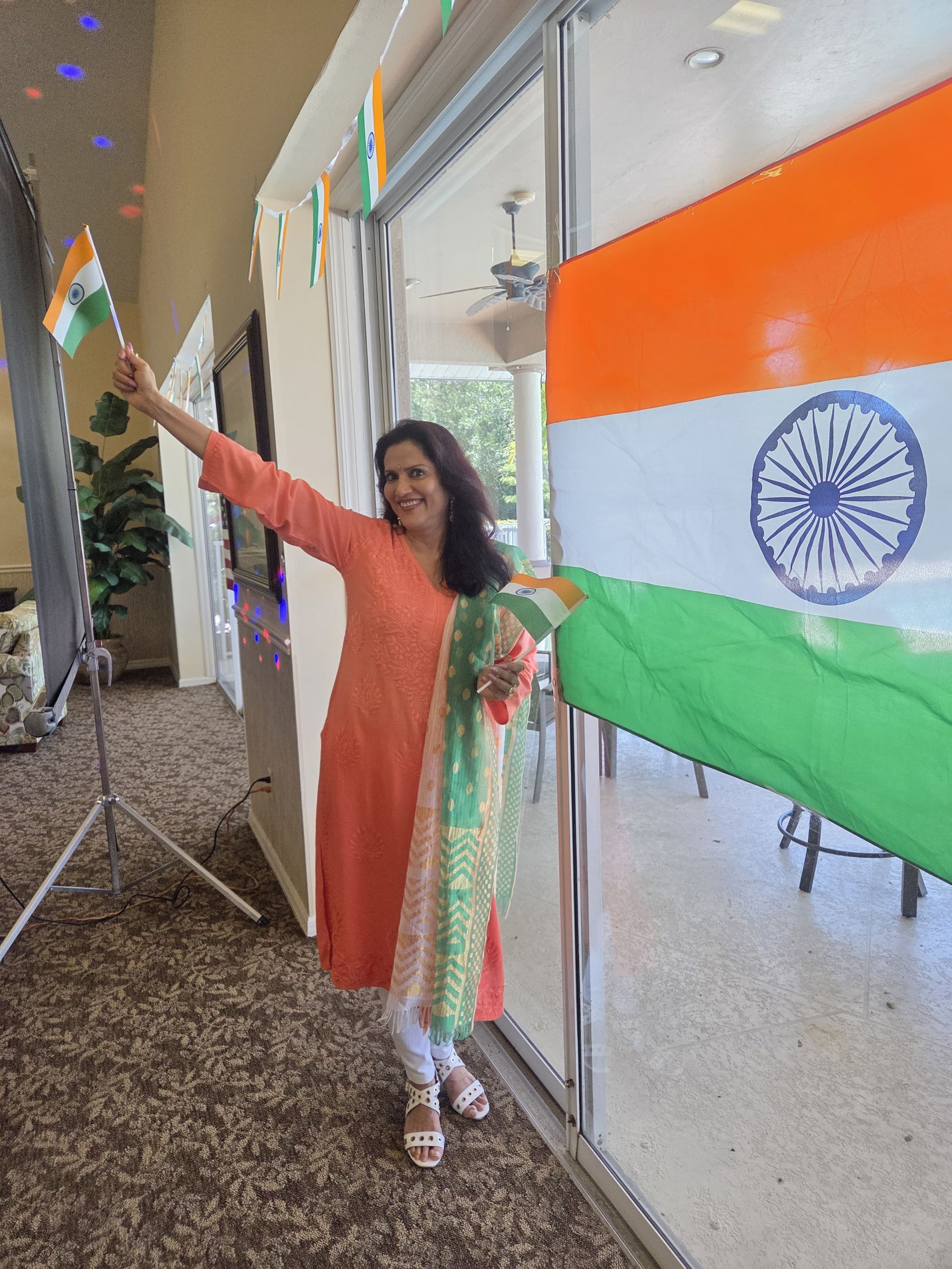 Woman in orange Indian suit waving an Indian flag, standing near a large flag in a decorated room.