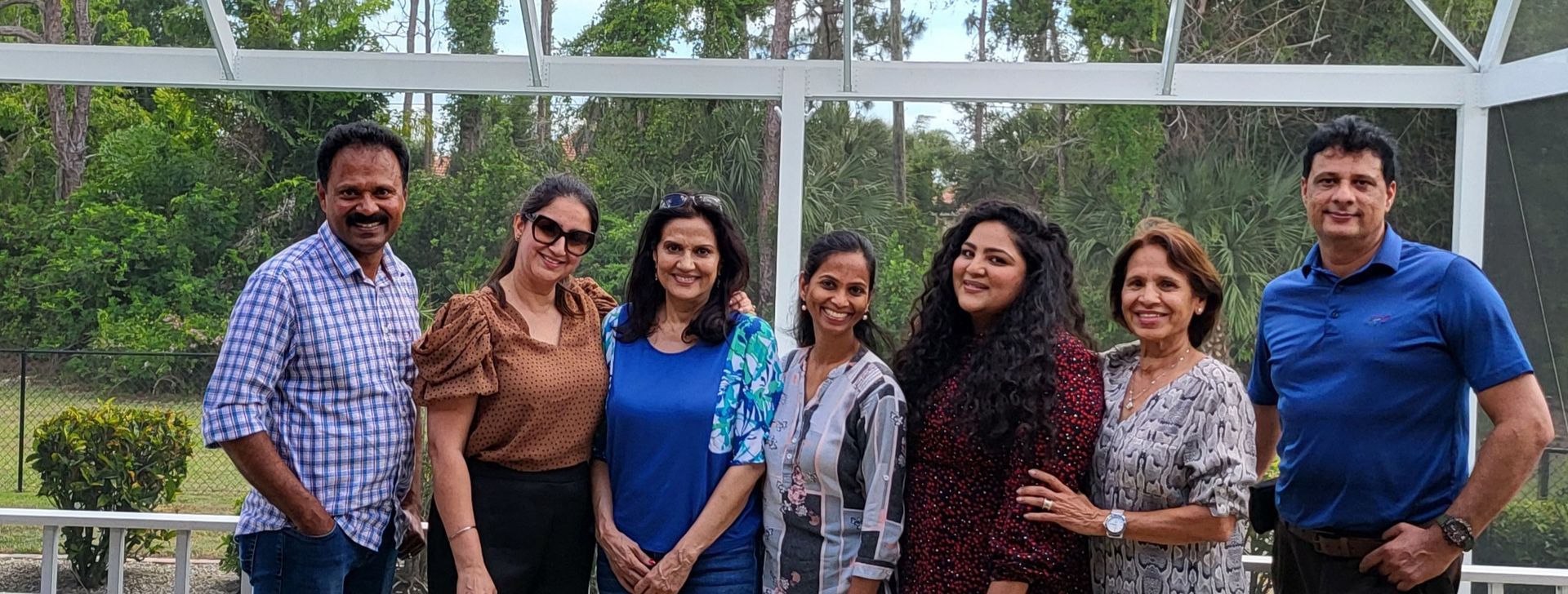 A group of seven people smiling outdoors. They are standing in front of a screen with trees in the background.
