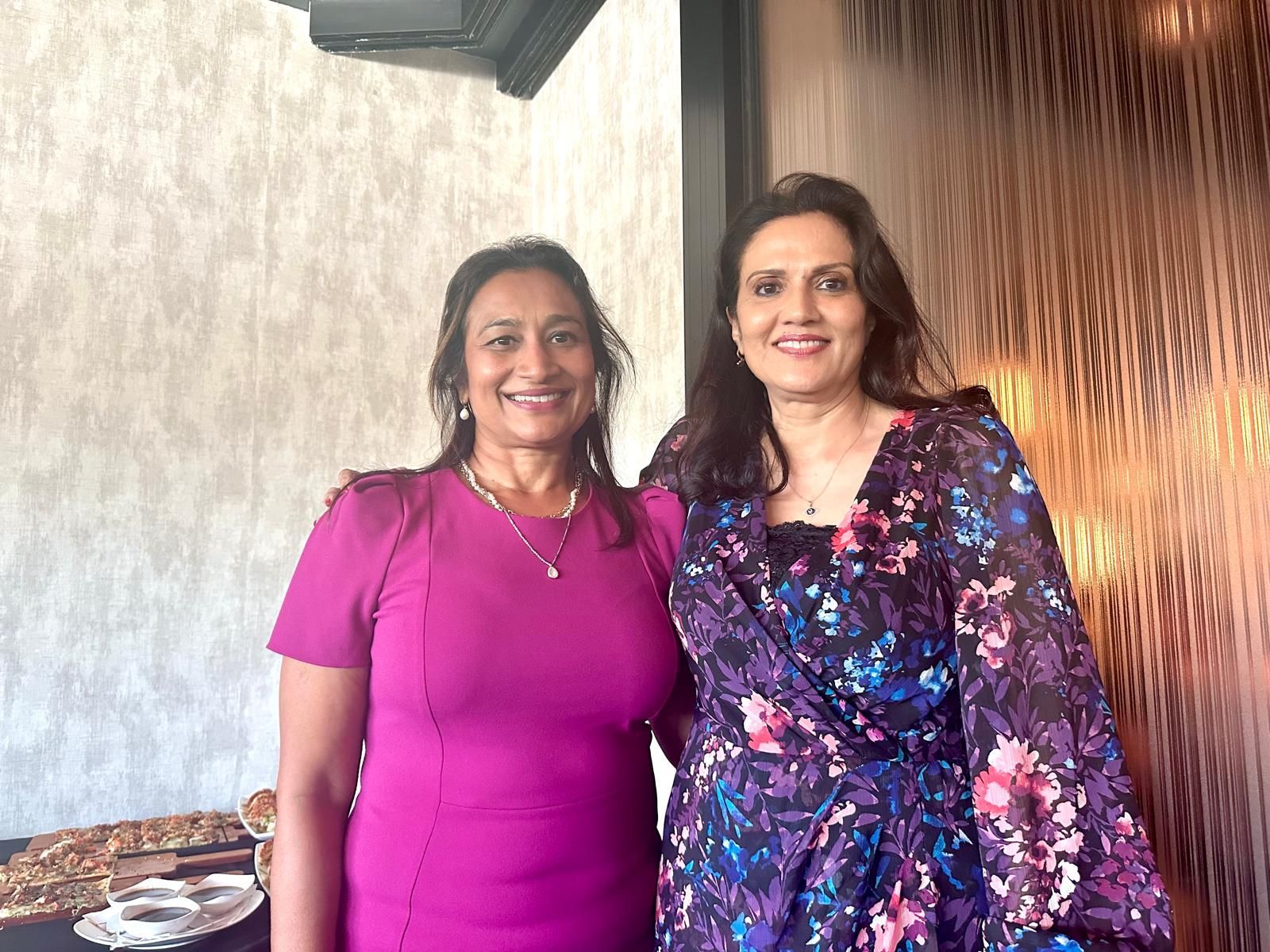 Two smiling women pose indoors. One in a pink dress, the other in a floral wrap dress.