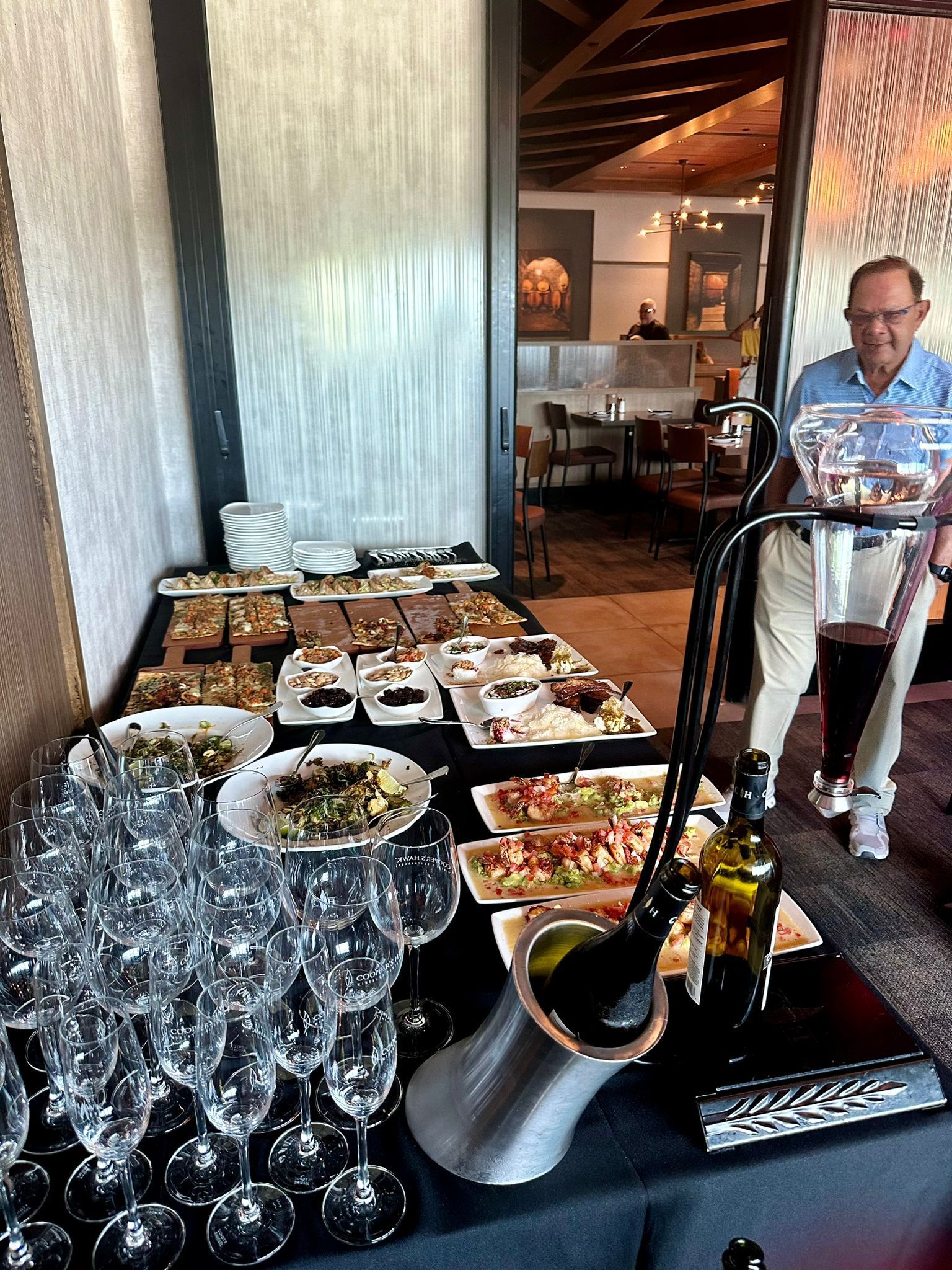 Buffet with food, glasses, and a server. A man carries wine to the table in a sunny room.