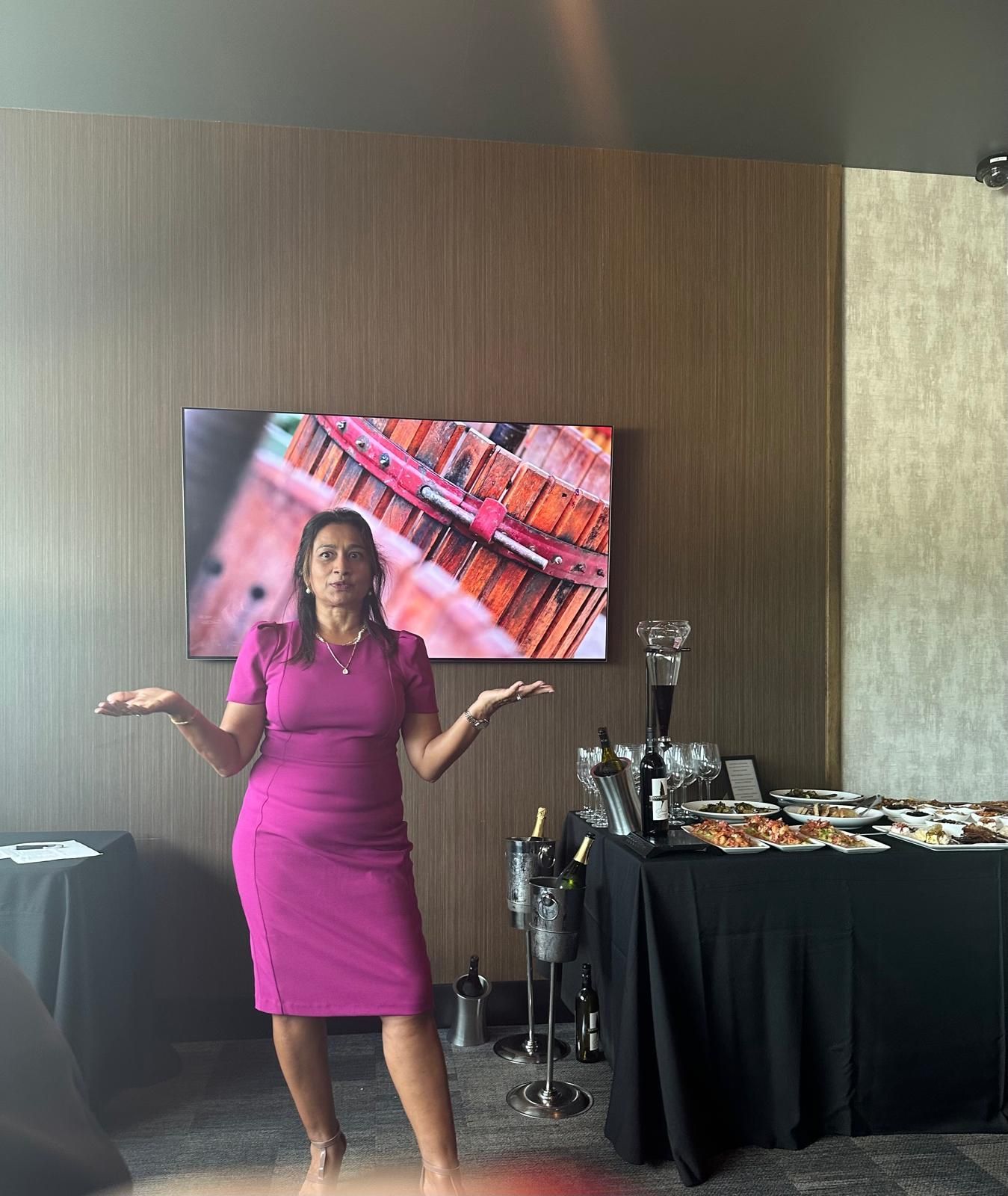 Woman in fuchsia dress gestures at catered food table. Setting appears to be an event with a TV screen and wine.