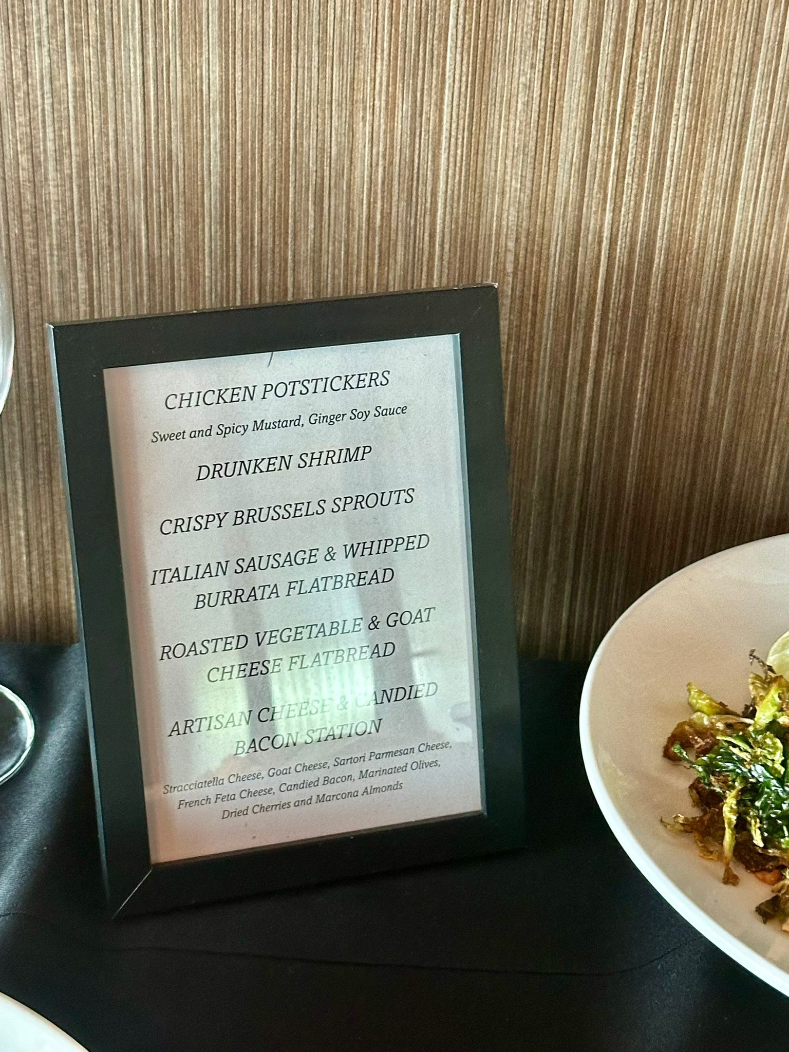 Menu in a black frame next to a white plate of food, set against a wood-paneled wall.