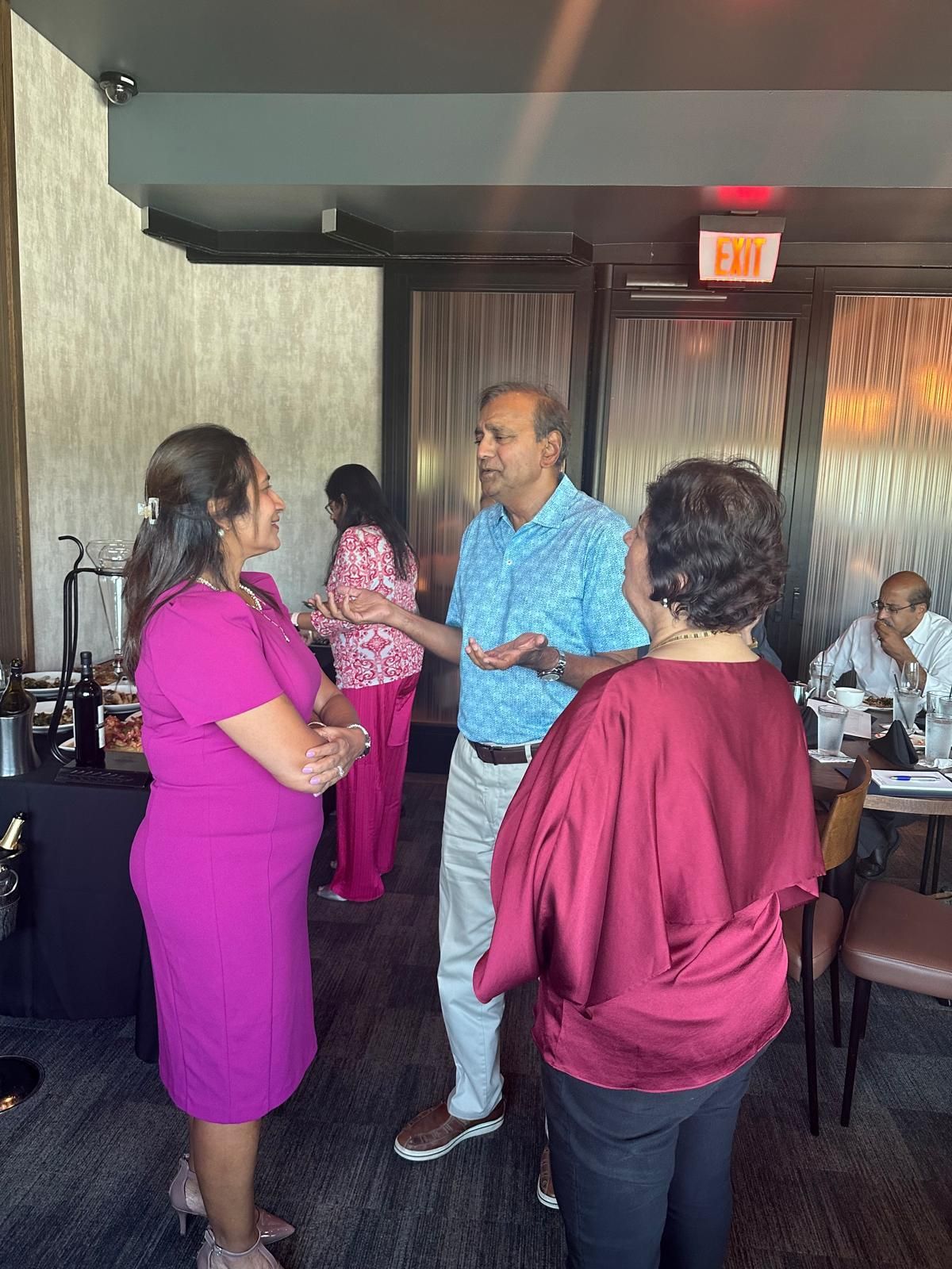 Group of people conversing in a restaurant. A woman in pink dress stands with another in a maroon shirt, while a man gestures.
