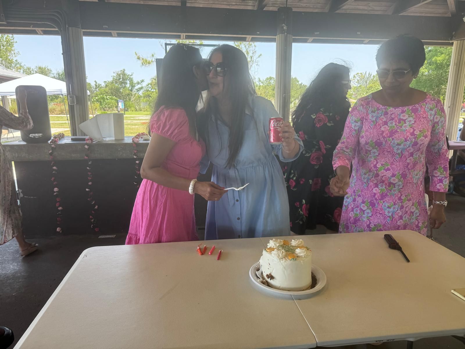 People at an outdoor party with a cake. Woman kisses another while another looks on.