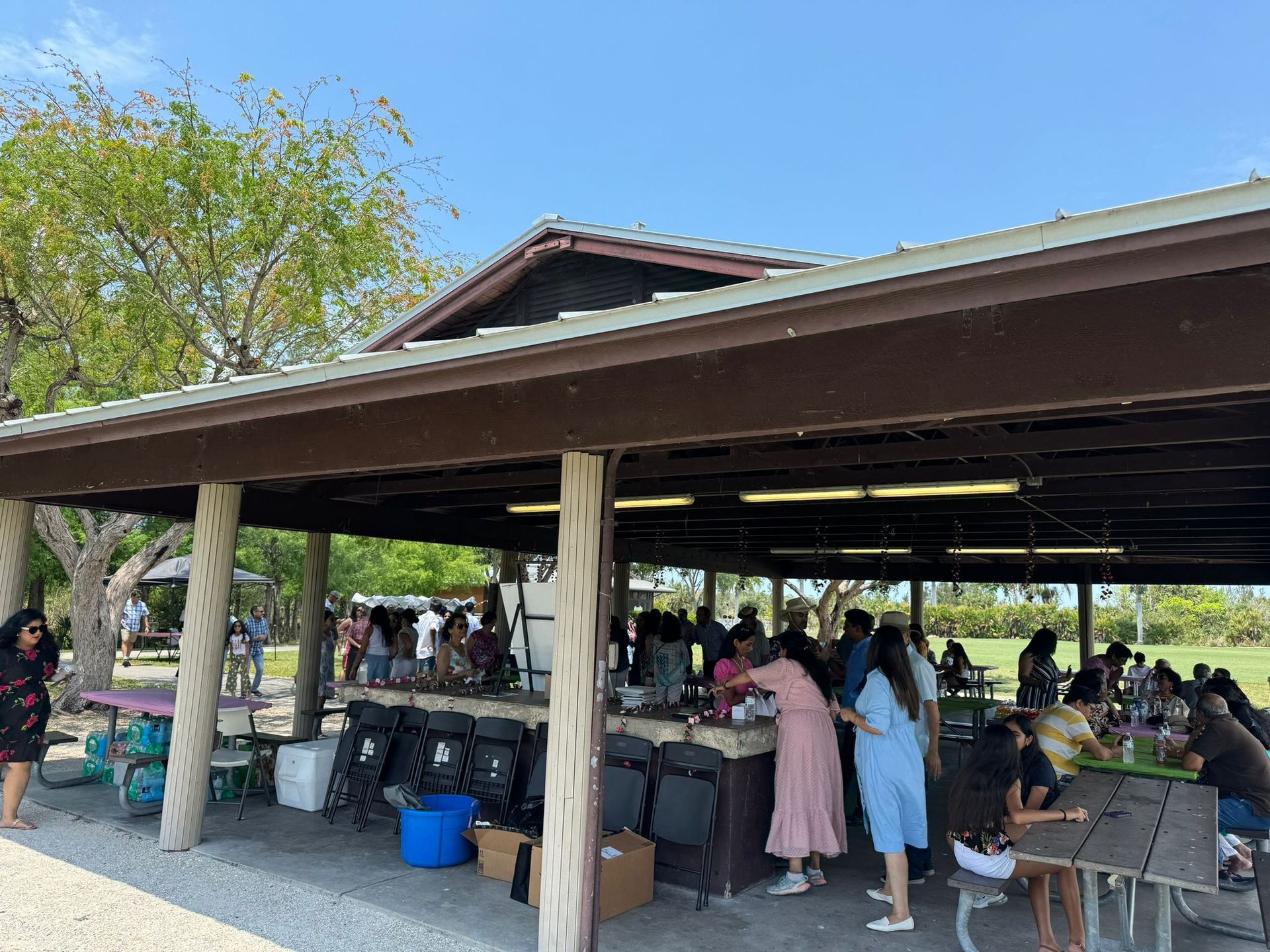 People gathered at a brown-roofed pavilion, preparing food and socializing on a sunny day.