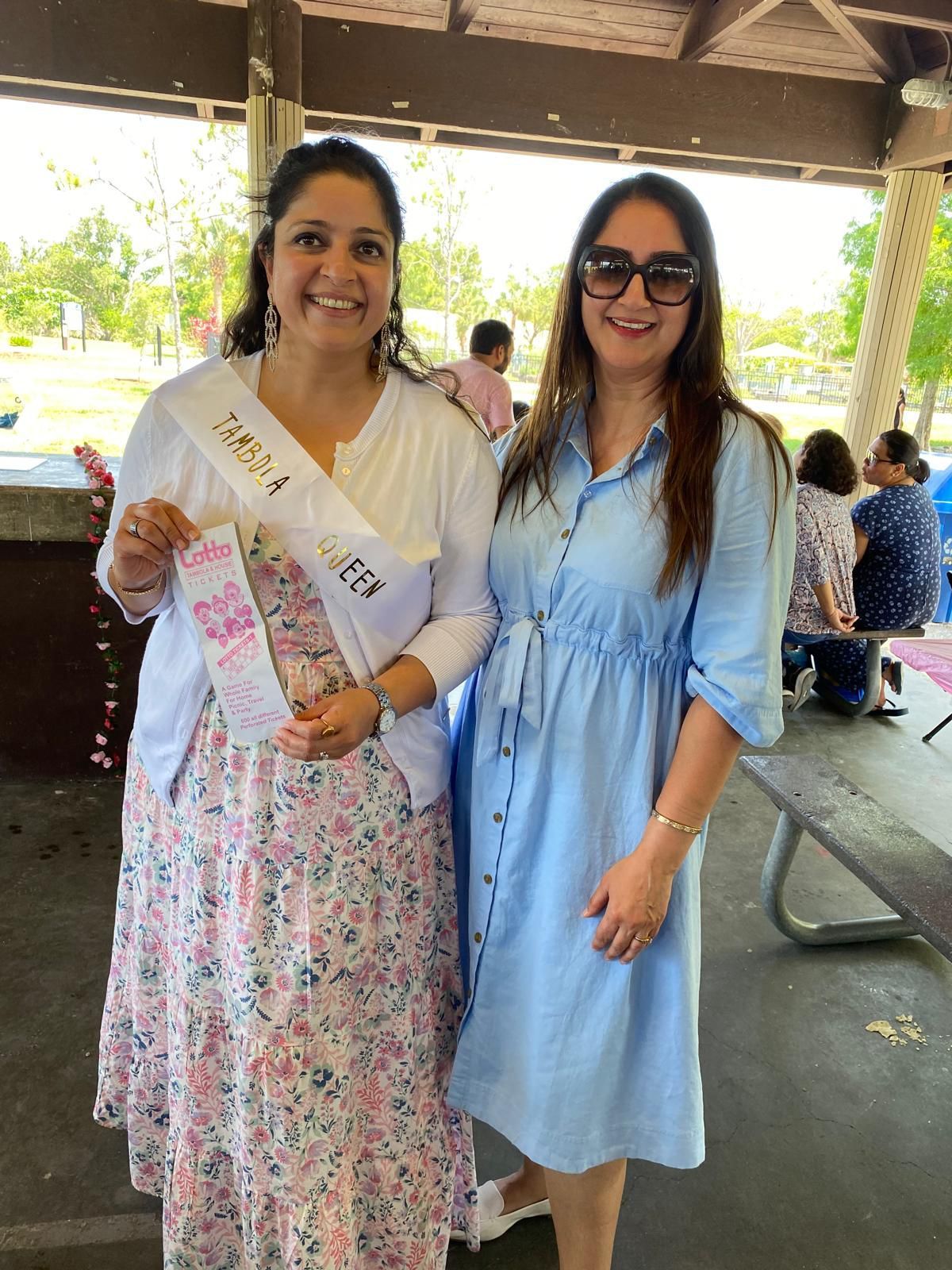 Two women smiling at an outdoor gathering. One wears a sash and floral dress; the other wears sunglasses and a blue dress.