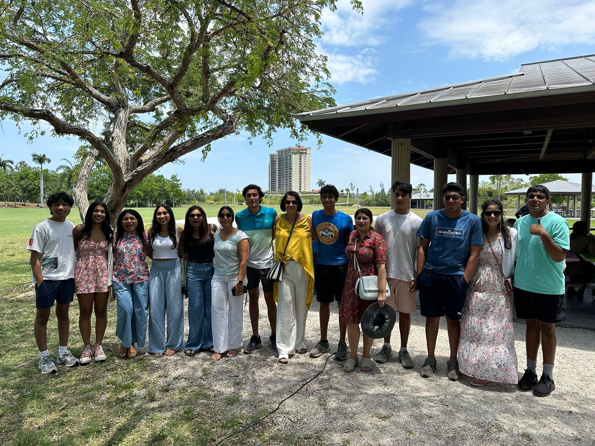 Group of people smiling for a photo at a park. Trees, shelter, and blue sky background.