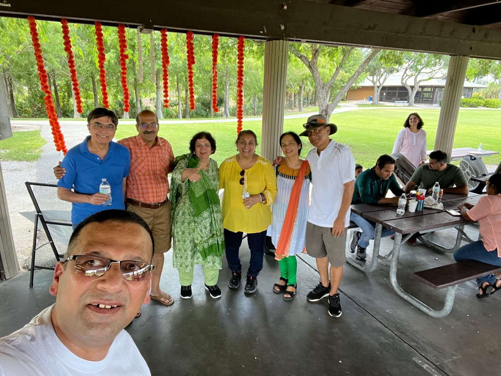Group of people at a picnic, smiling. Some are wearing colorful clothes, outside under a covered area with decorations.