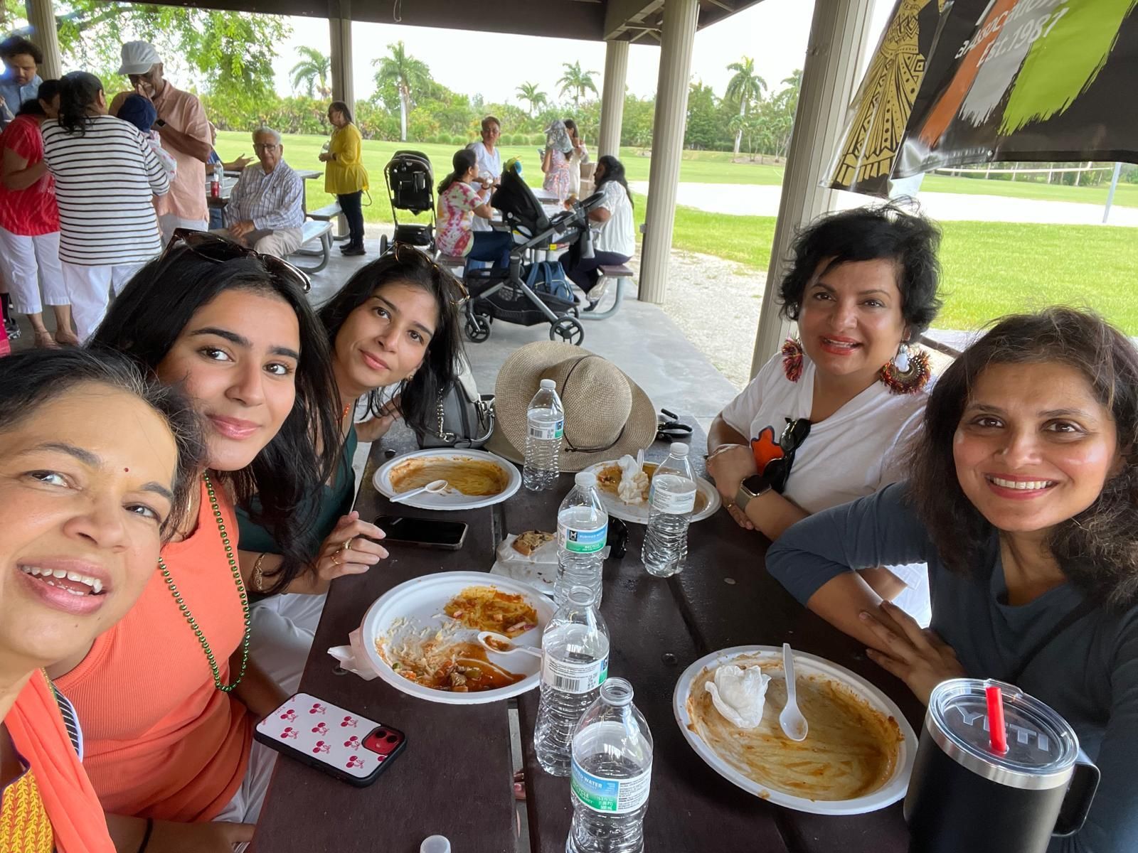 A group of women smile at a picnic table with food and drinks outdoors under a shelter.