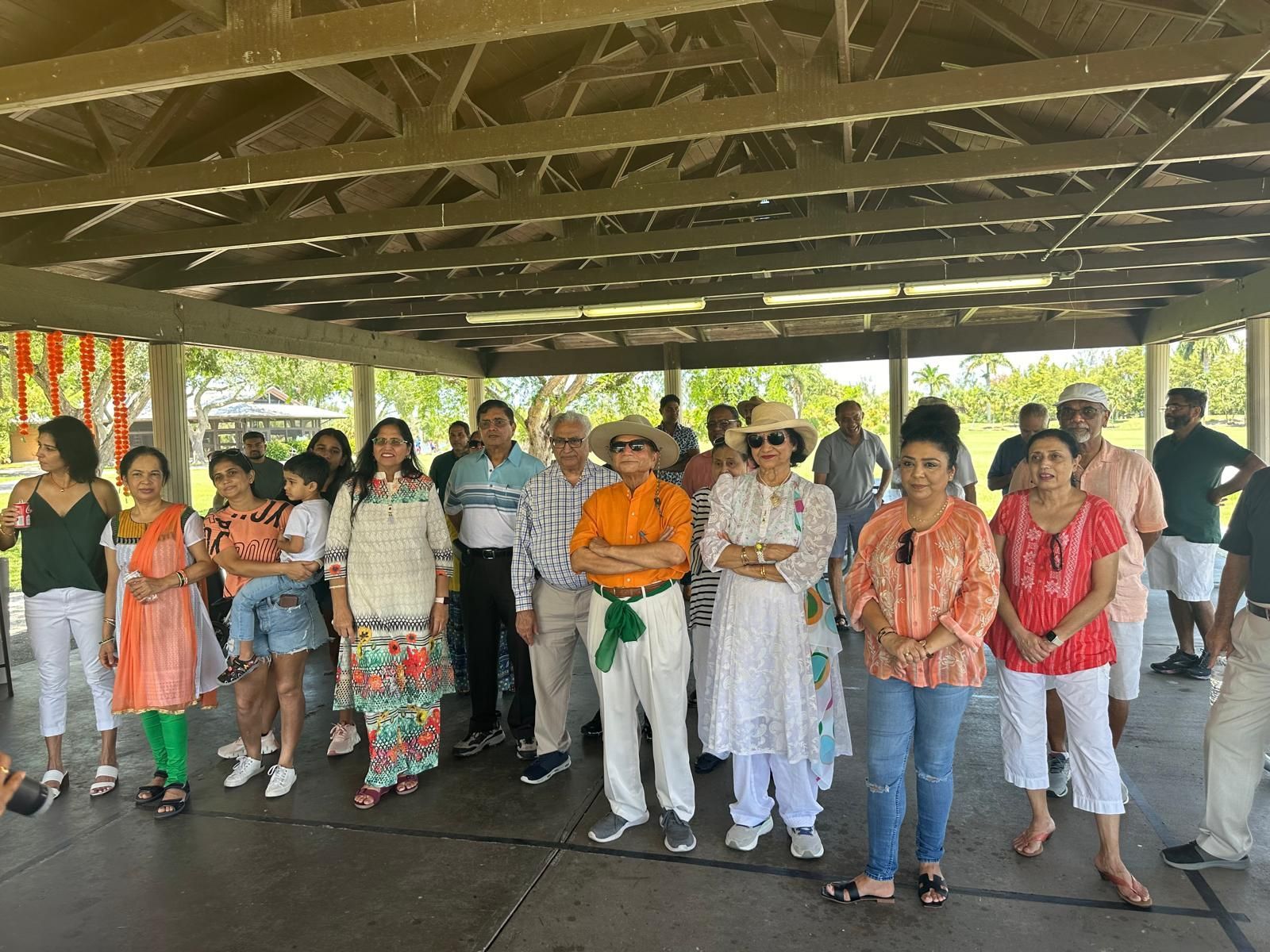 Group of people posing under a shelter; many wearing colorful attire, possibly at an outdoor event.