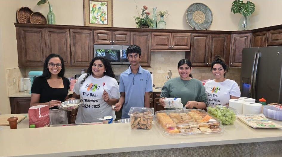 Five people in a kitchen with food items; smiling and preparing meals.
