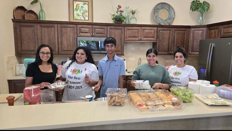 Five people in a kitchen with food. Two women in matching shirts smile, gesturing. Others hold snacks.